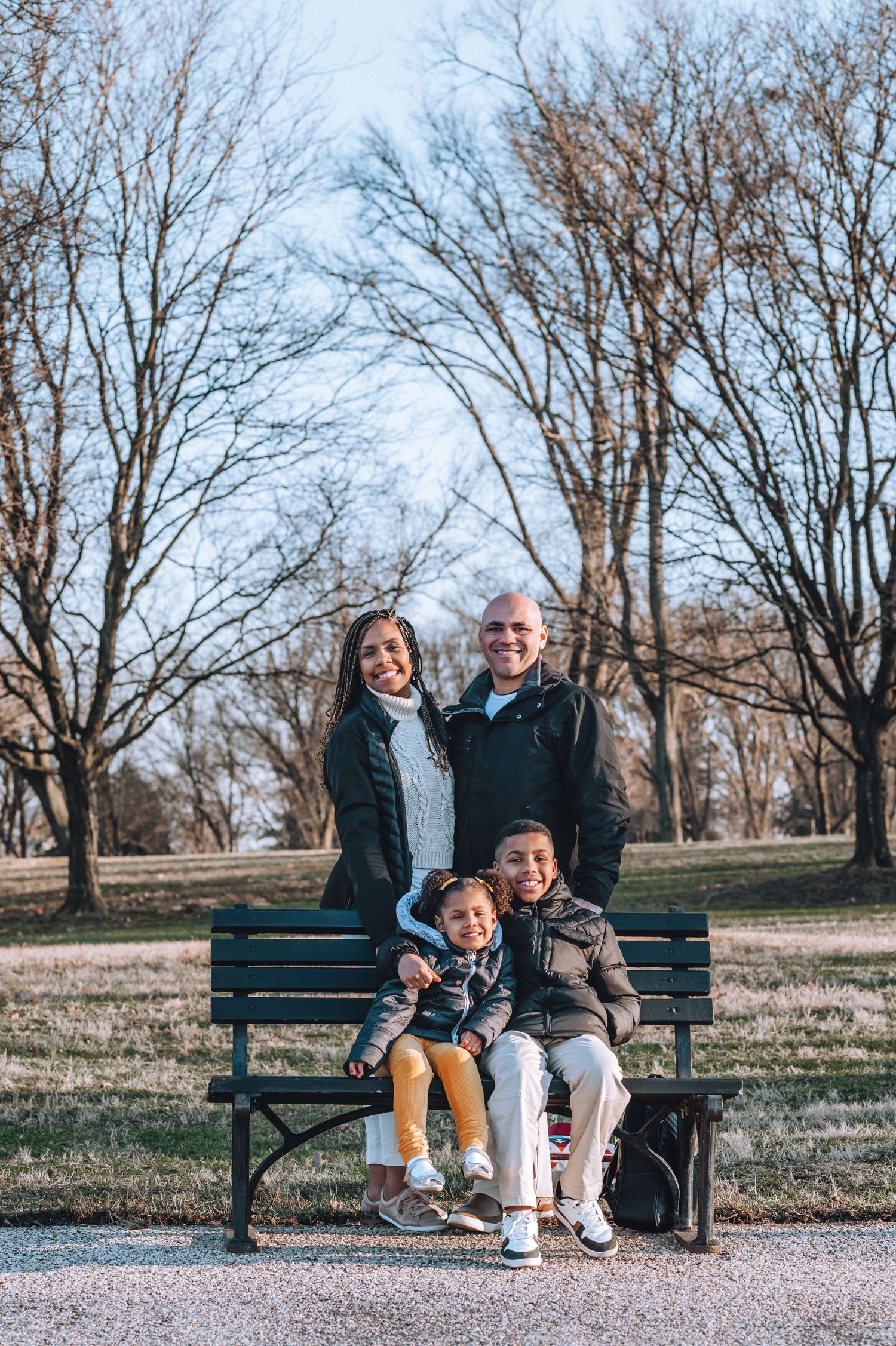 A family sits together on a green park bench surrounded by bare winter trees on a sunny day.