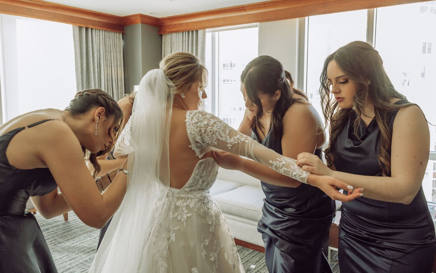 Three people in dark dresses help adjust a beautiful lace wedding gown in a sunlit room.
