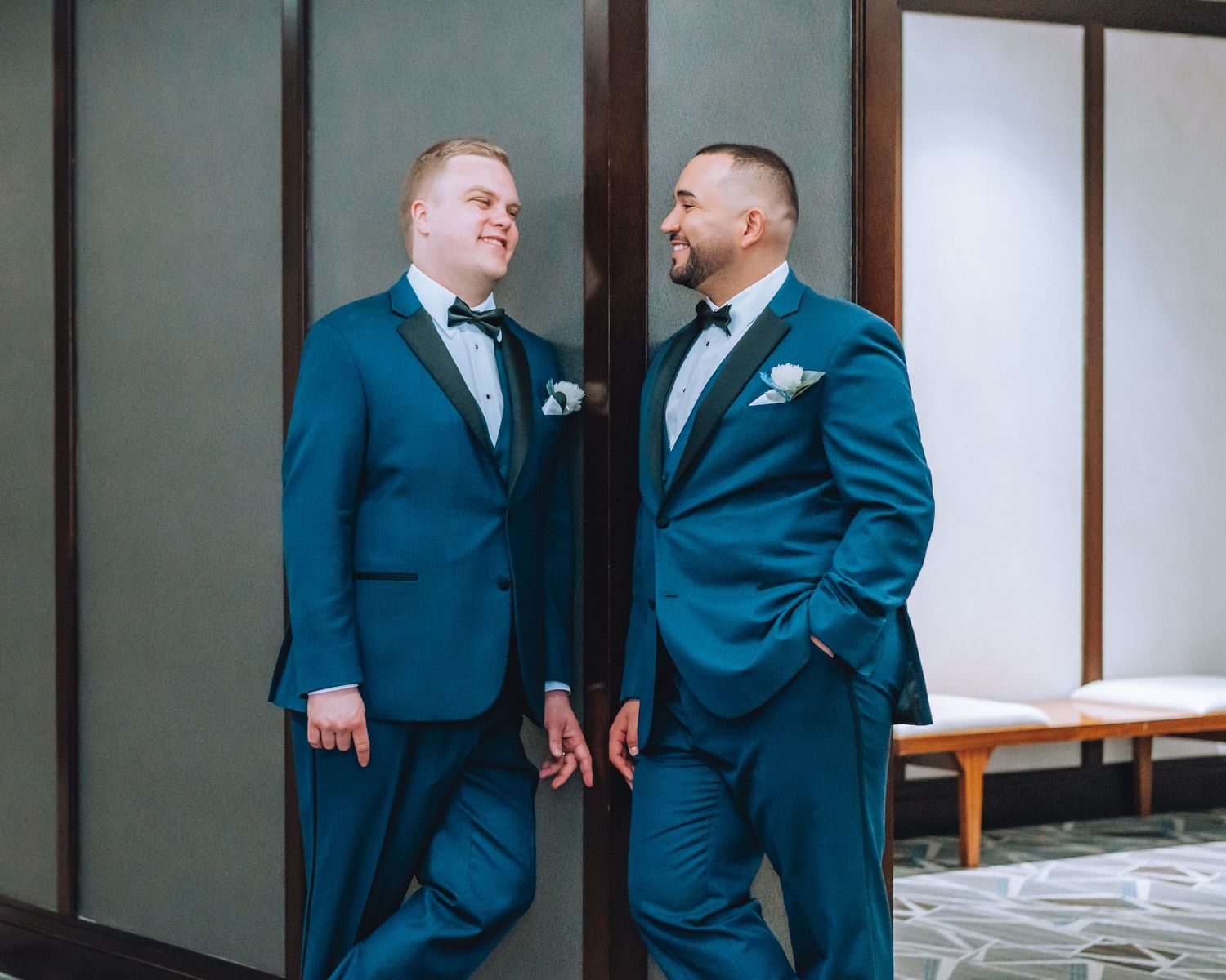 Two people wearing matching navy blue tuxedos with bow ties laugh together in a hallway.