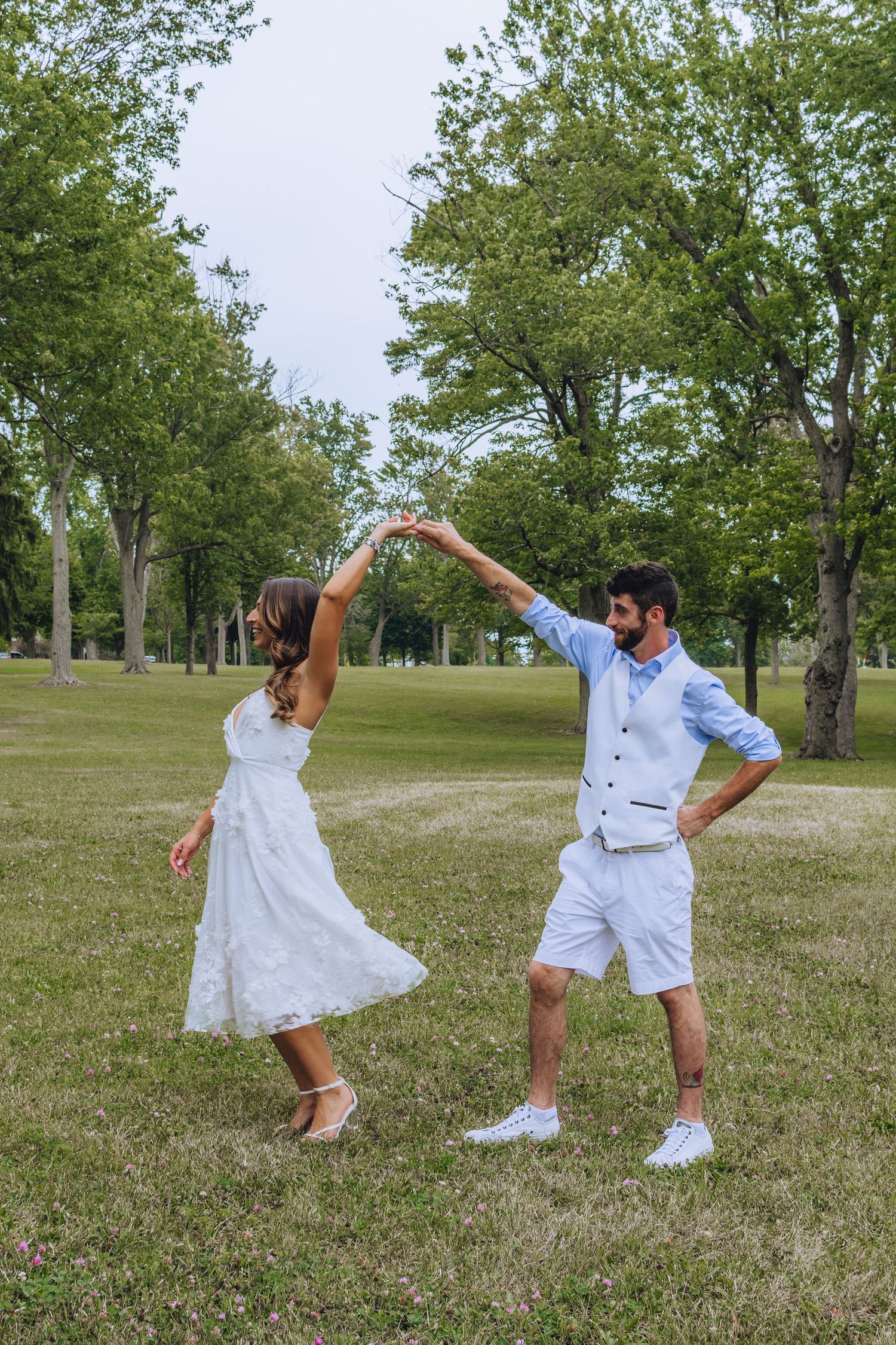 Two people in white outfits dance playfully together on a grassy hill with trees in the background.