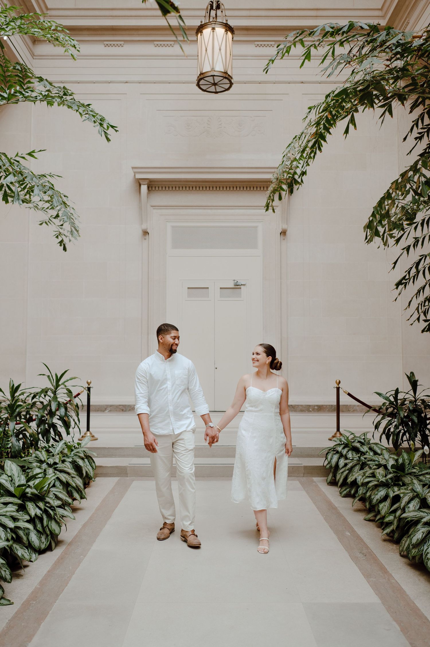 A couple in coordinating white outfits playfully walks through an indoor garden hallway.