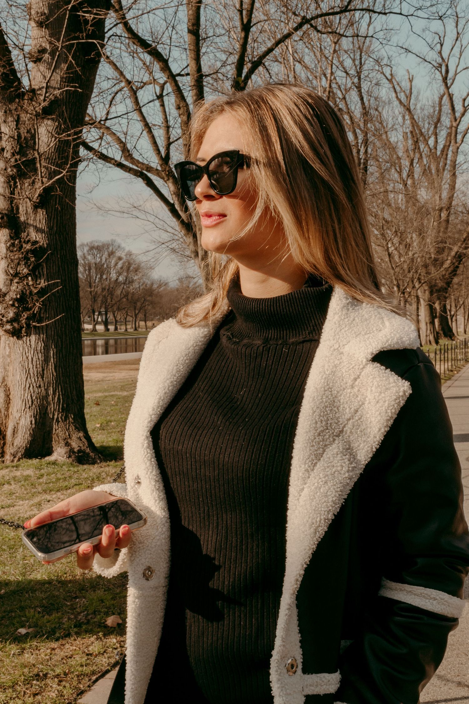 Woman in black turtleneck and white coat smiles while wearing oversized sunglasses outdoors.