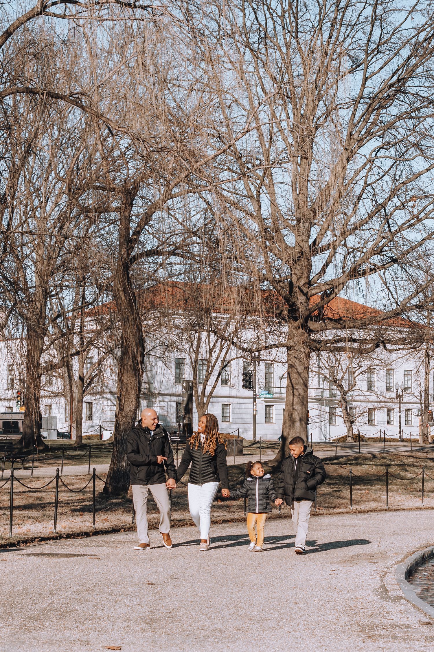 Family walks together on path with historic architecture visible through winter trees.