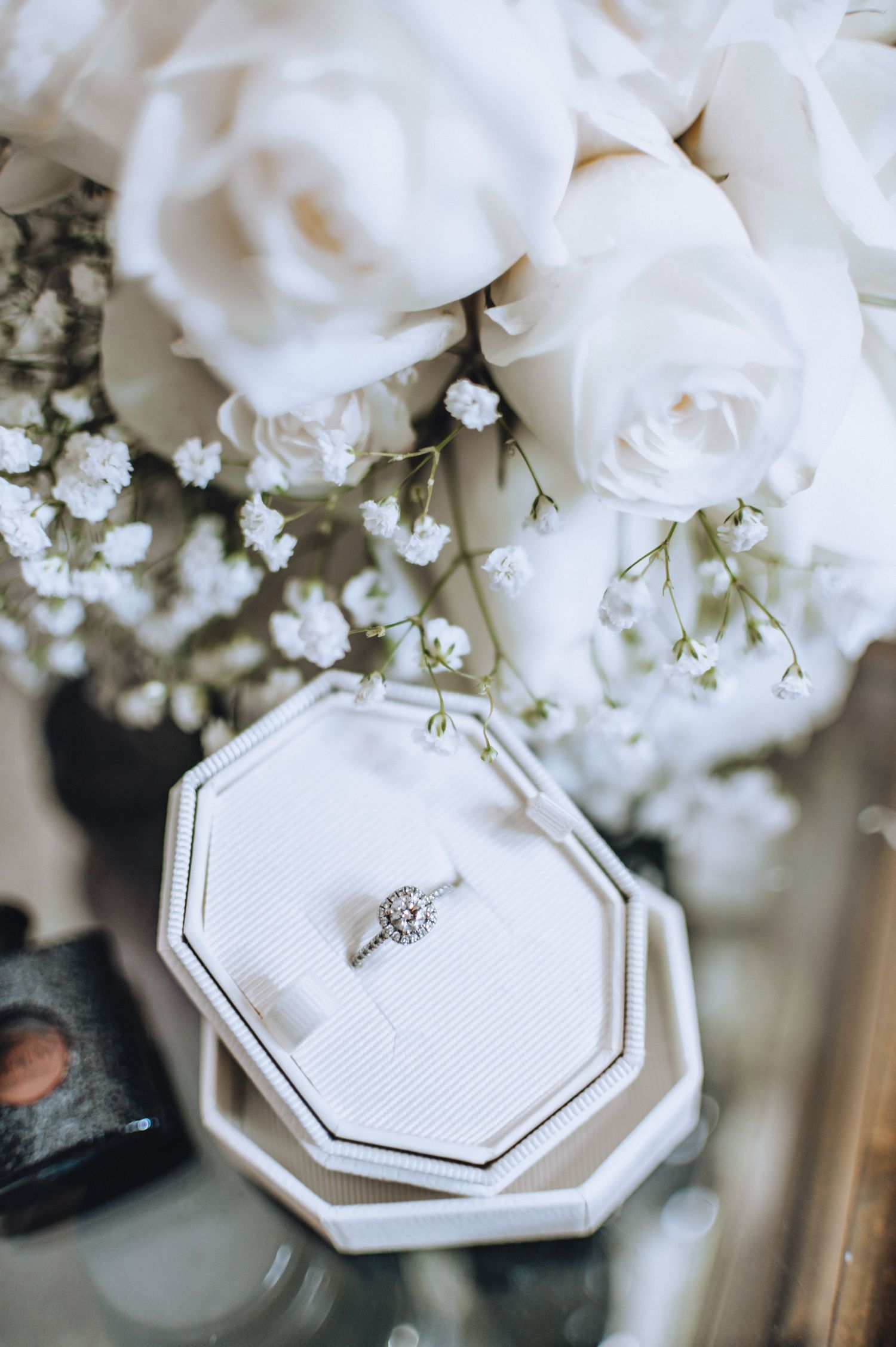Elegant engagement ring on white octagonal ring box surrounded by white roses and baby's breath flowers.