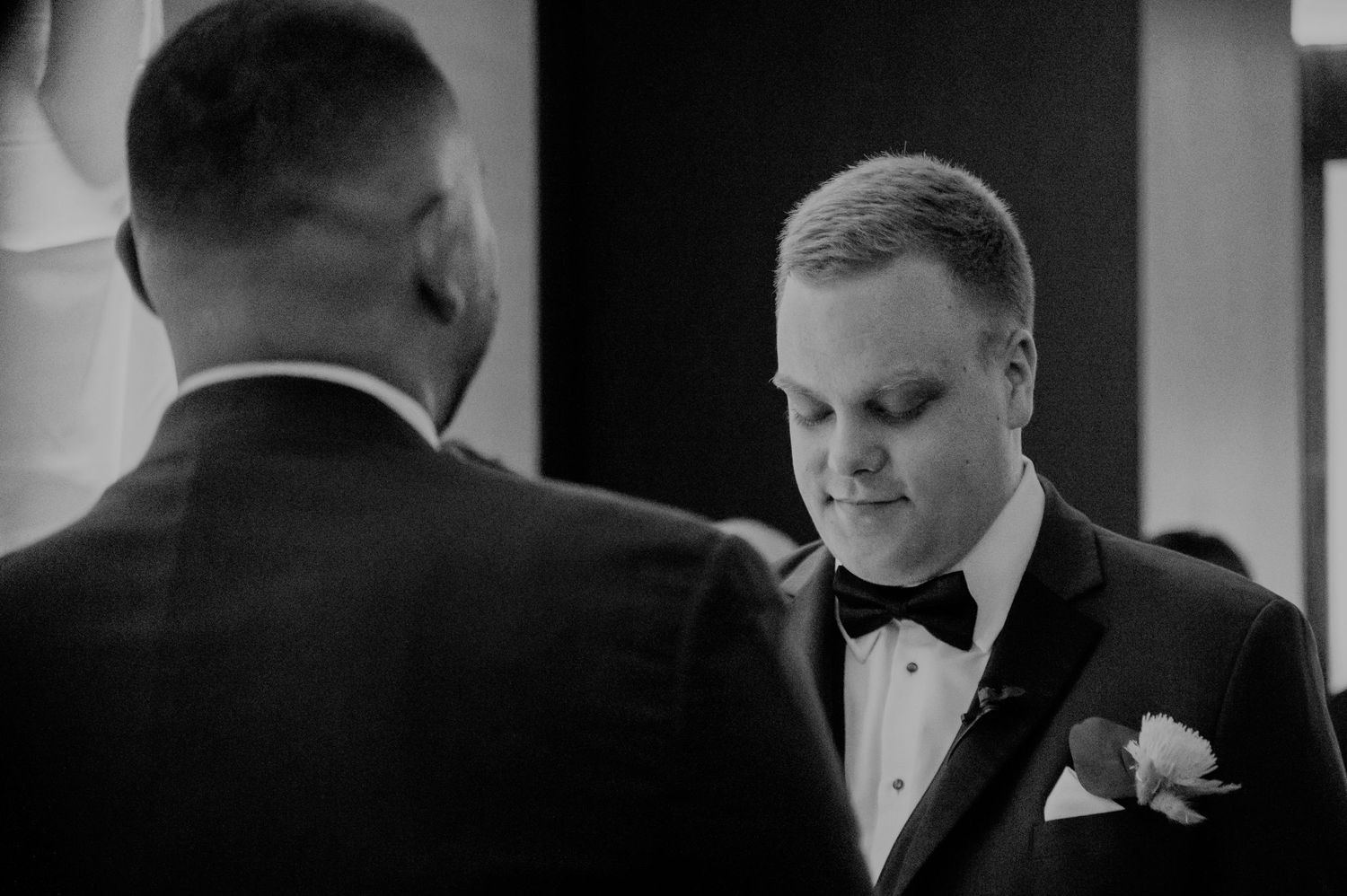 A black and white photograph captures a formal moment during a wedding ceremony as a guest in a tuxedo looks downward.