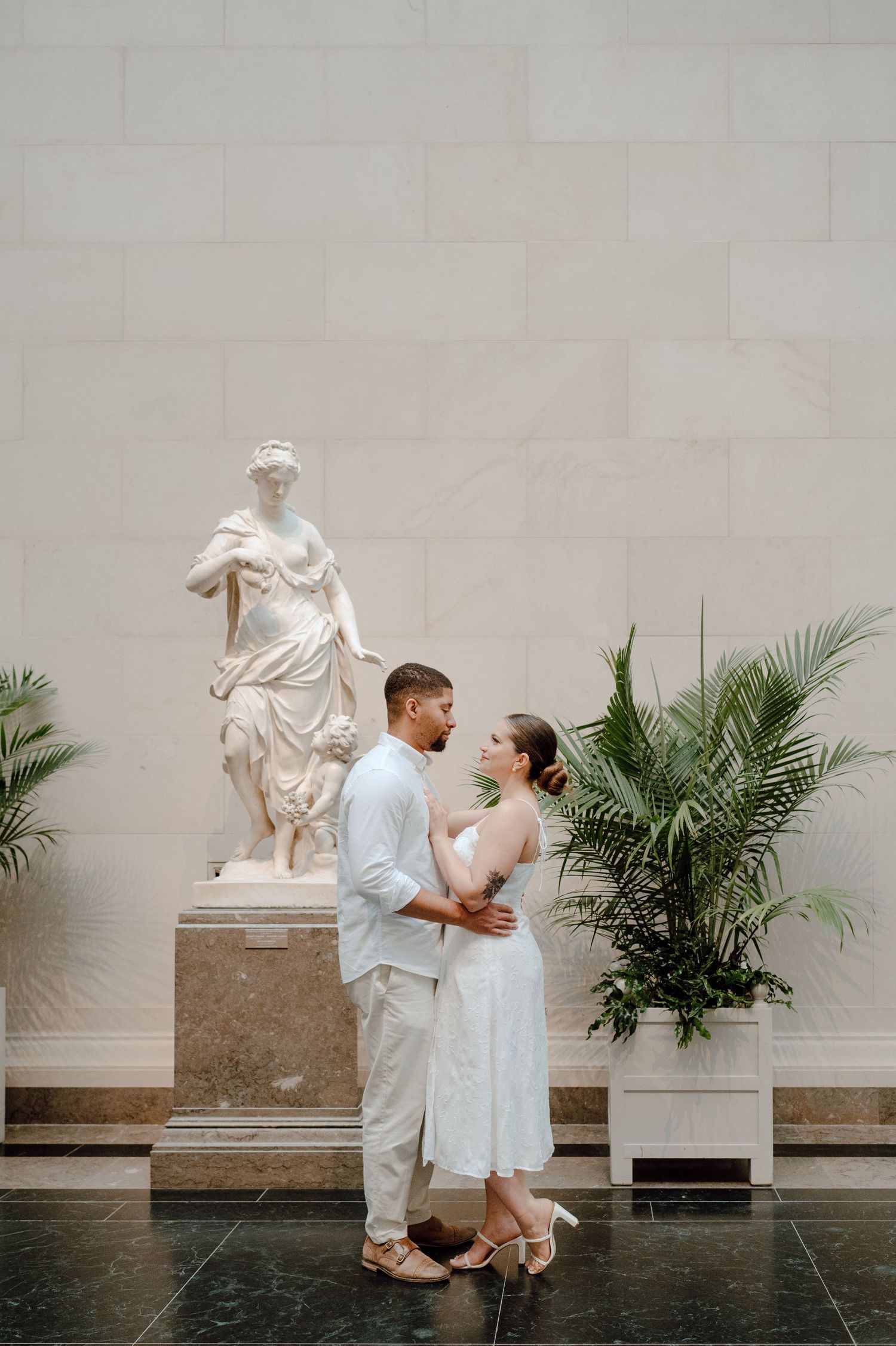 A couple shares an intimate moment in an elegant museum gallery with a classical statue and palm plants.