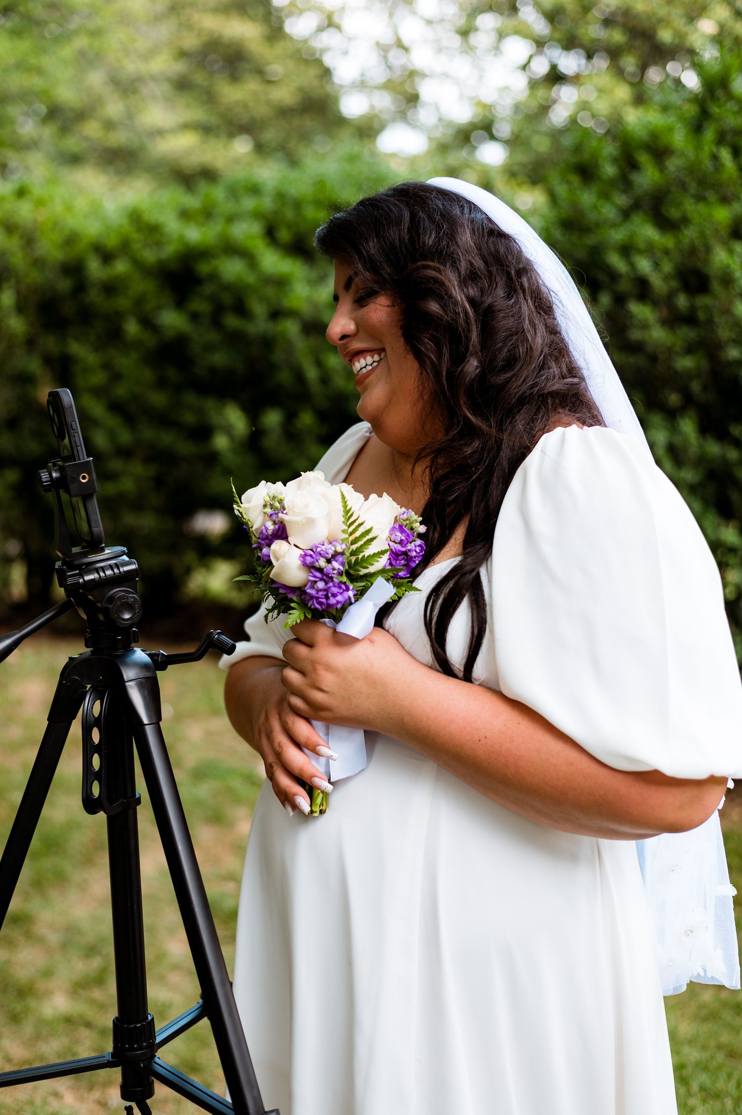 A joyful bride in a white dress holds a purple bouquet while standing next to a camera tripod outdoors.