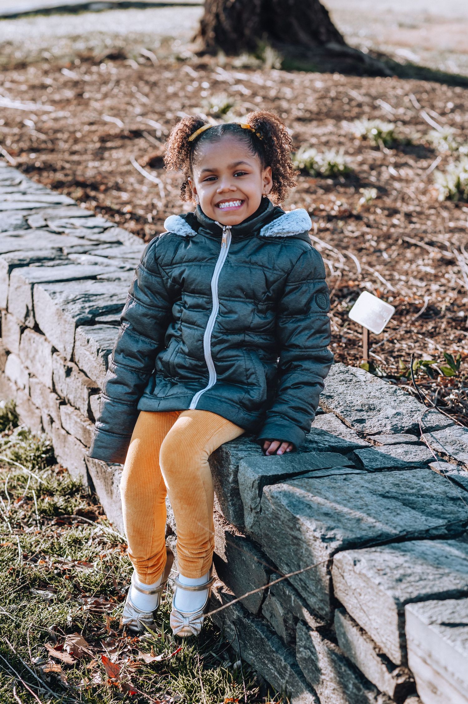 Child in teal winter coat and yellow pants sits on stone wall outdoors.