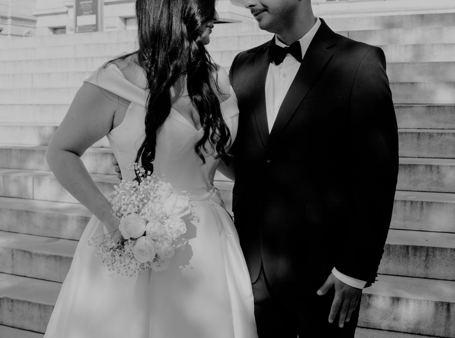 Black and white photo of a bride holding a bouquet standing next to her groom in formal wear.