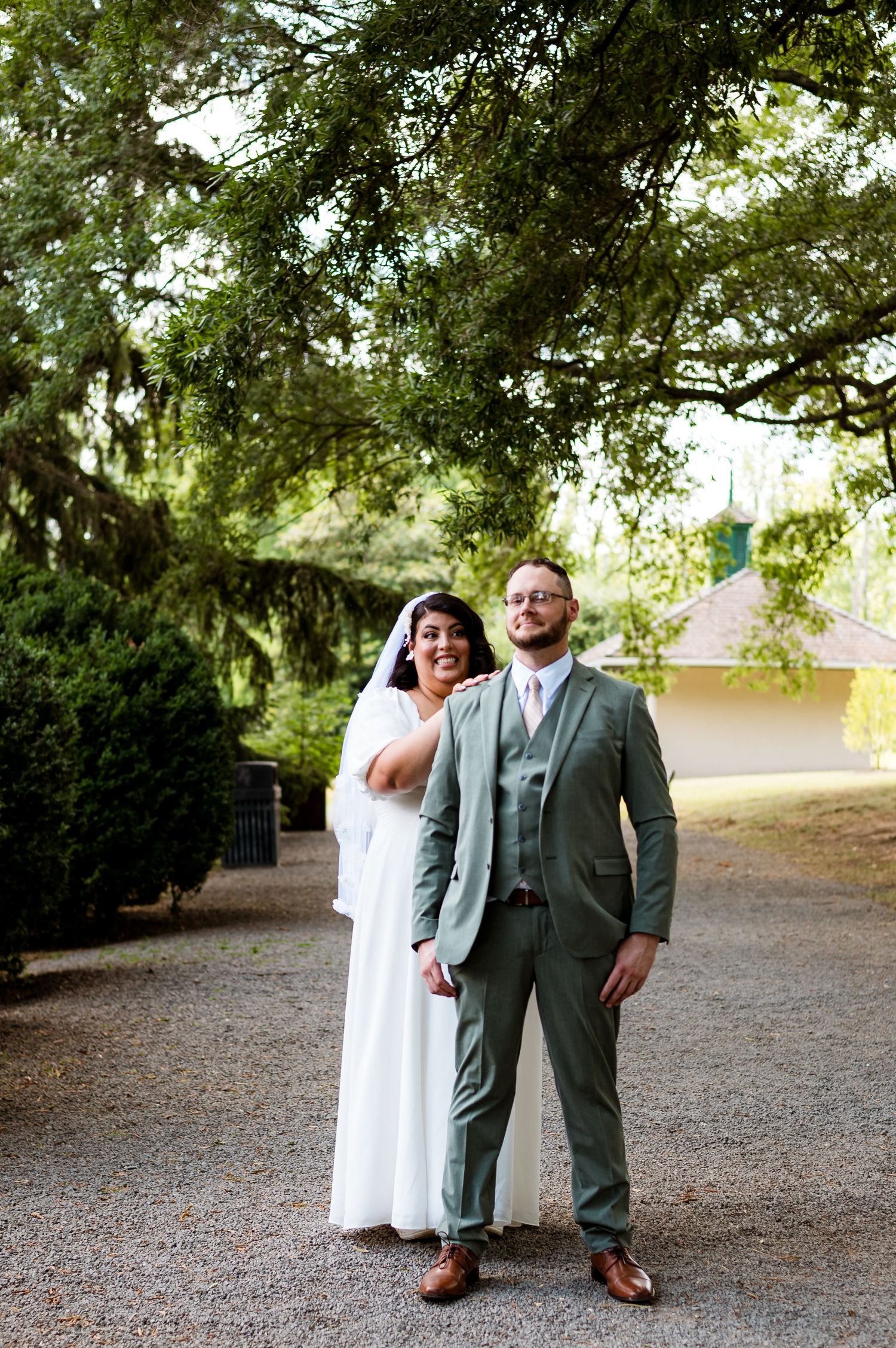 A couple in wedding attire poses together on a gravel path under lush green trees on their special day.