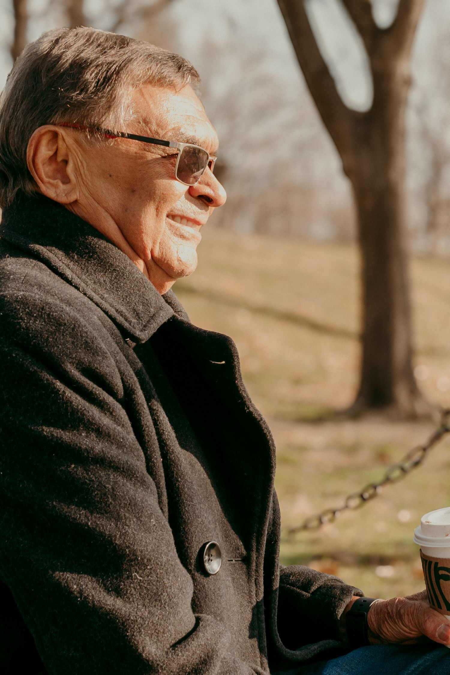Person in winter coat smiles while holding coffee outdoors on sunny day.