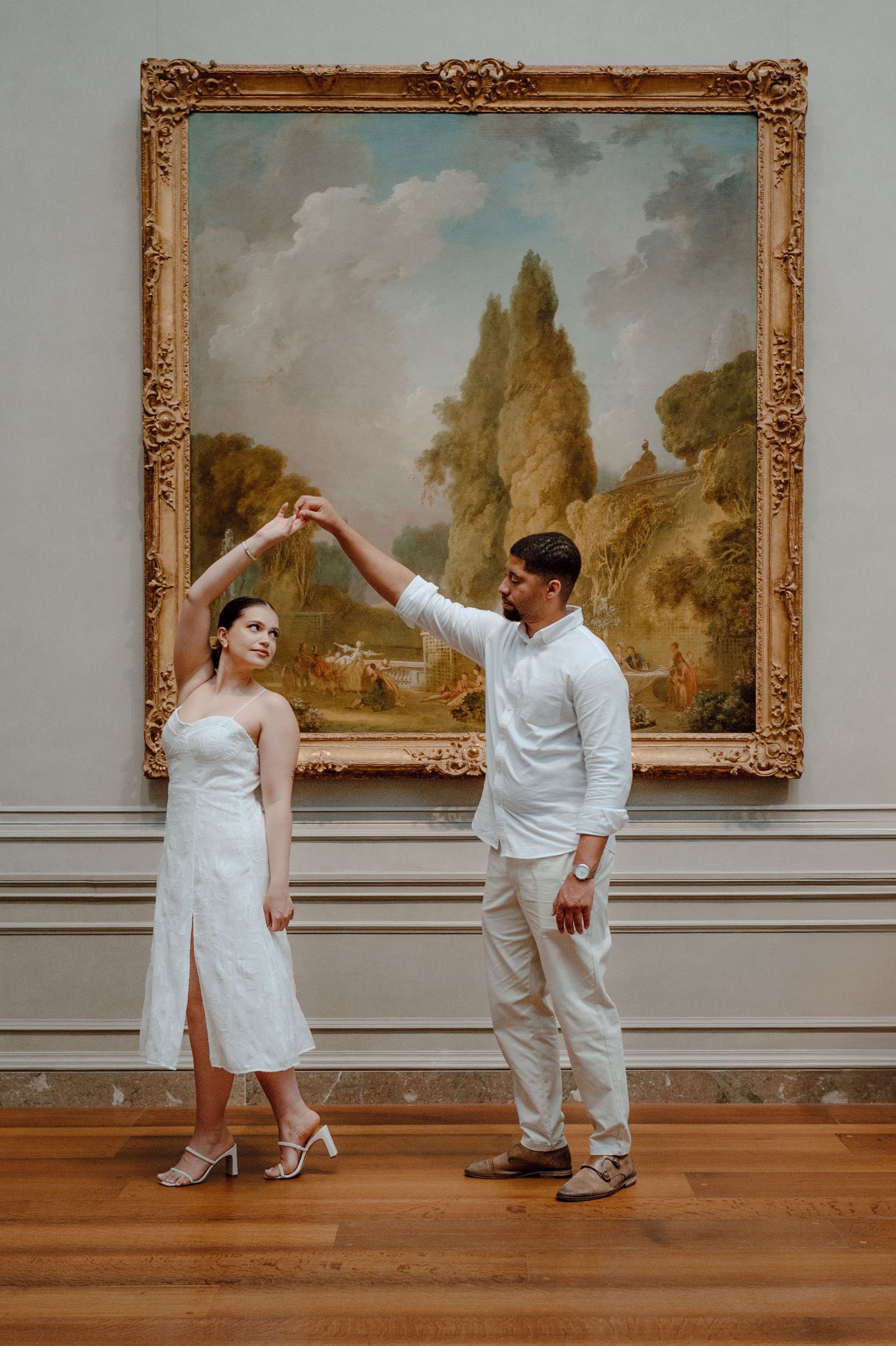 Two people in elegant white outfits strike a dance pose in front of a large ornate framed landscape painting in a museum.