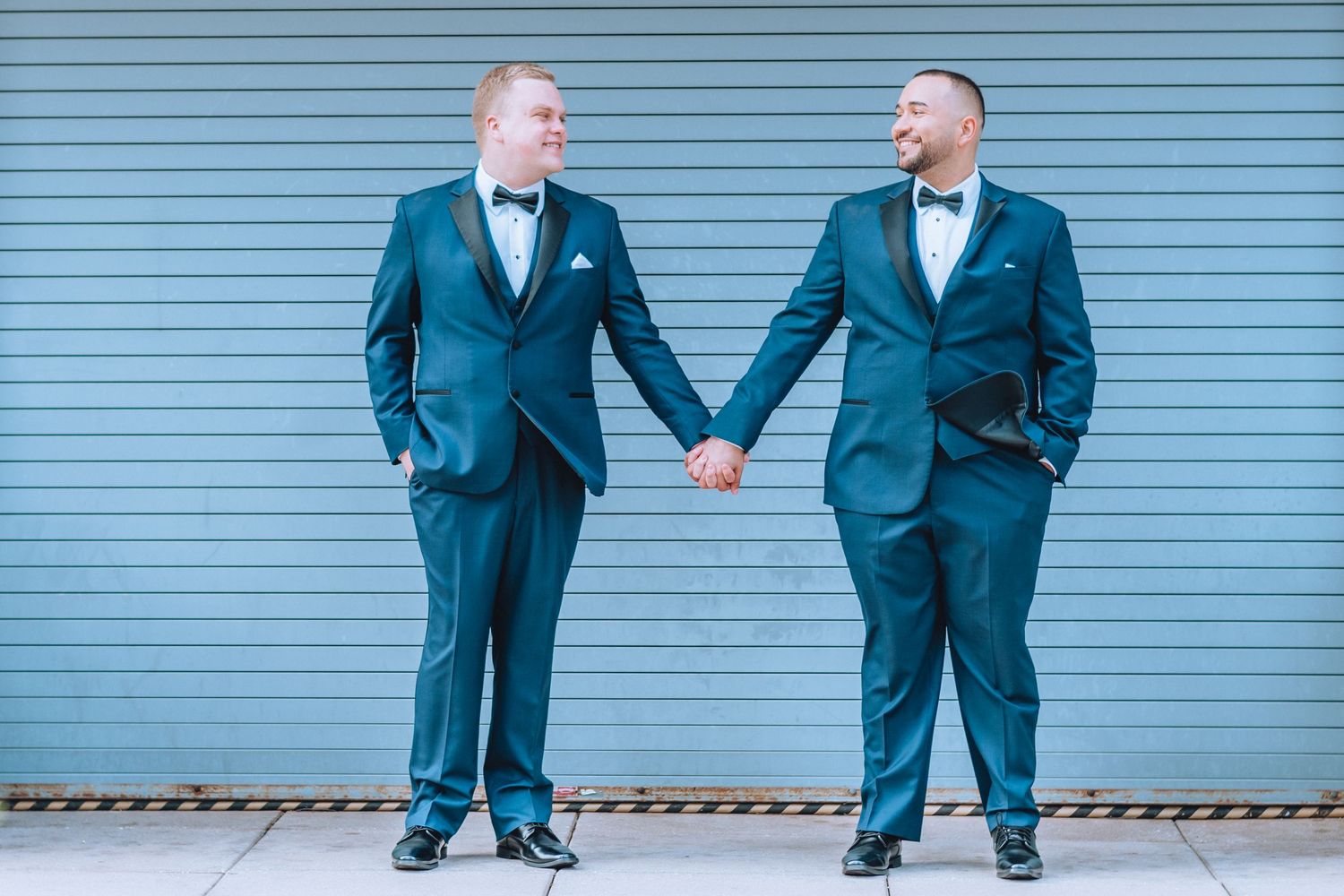 Two grooms in matching navy blue tuxedos hold hands while standing against a light blue corrugated wall.