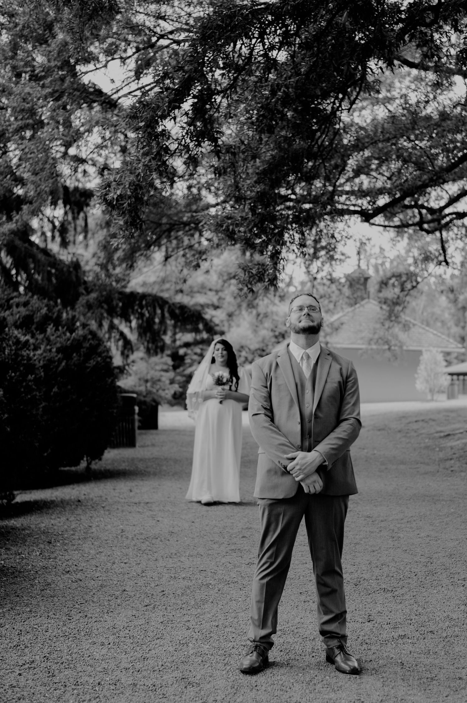 Black and white photo of a wedding couple standing apart in a garden setting under large trees.