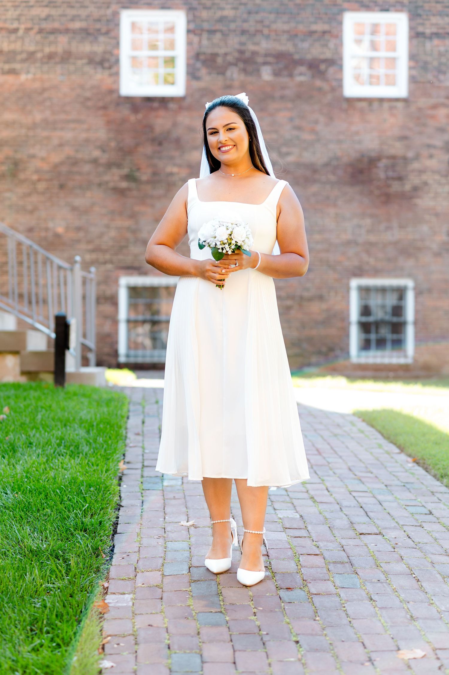 A bride in a tea-length white dress stands on a brick pathway holding a small bouquet against a stone building.