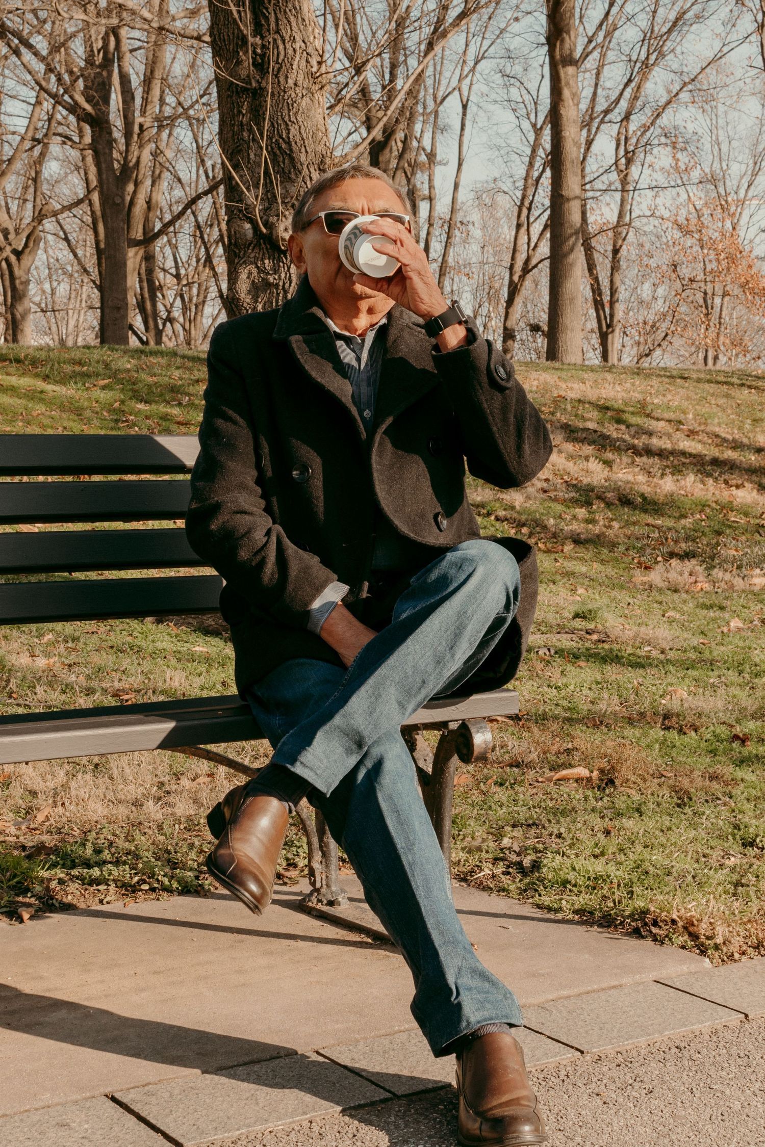 Person in dark coat sits on park bench drinking coffee on autumn morning.