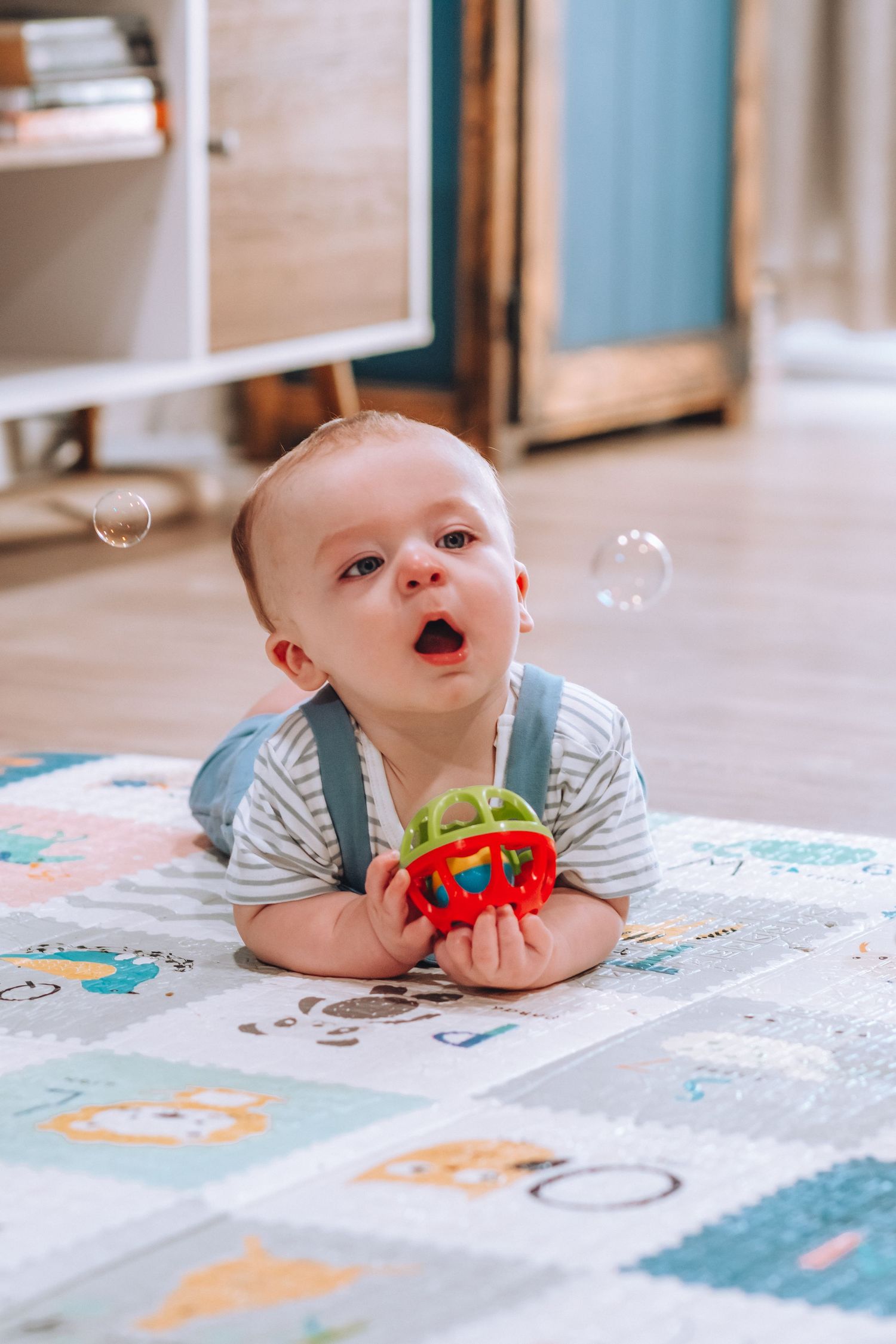 Baby on playmat watching bubbles while holding red and green toy.
