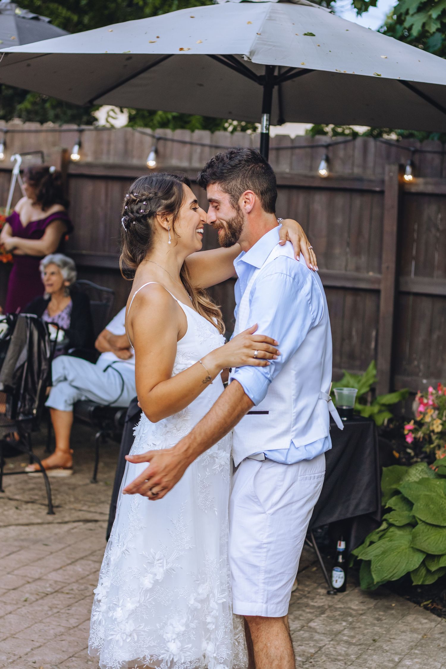 A couple embraces during a backyard wedding reception under an umbrella with string lights and greenery in background.