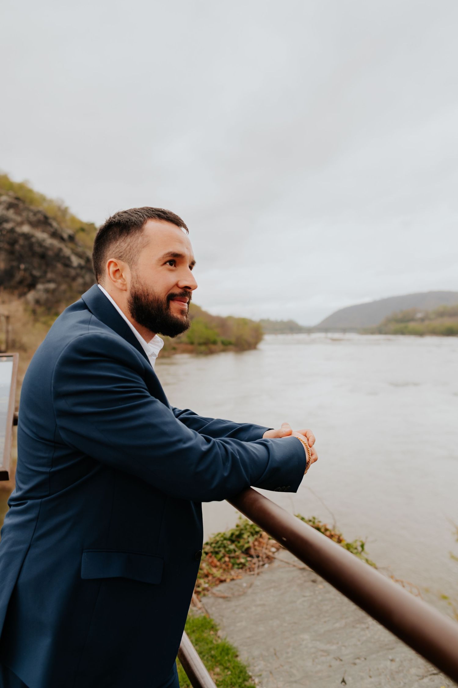 A person in a navy suit smiles while leaning against a railing overlooking a scenic river and mountains.