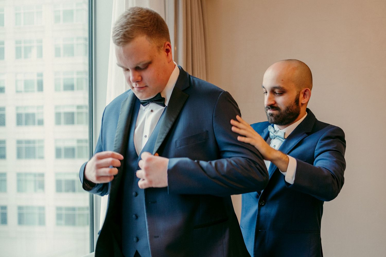 Two men in navy suits prepare formal attire by a large window in an elegant setting.