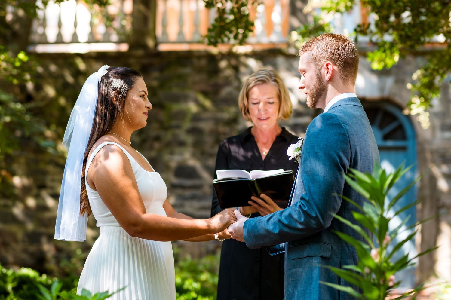A bride and groom hold hands during their outdoor garden wedding ceremony with an officiant presiding.