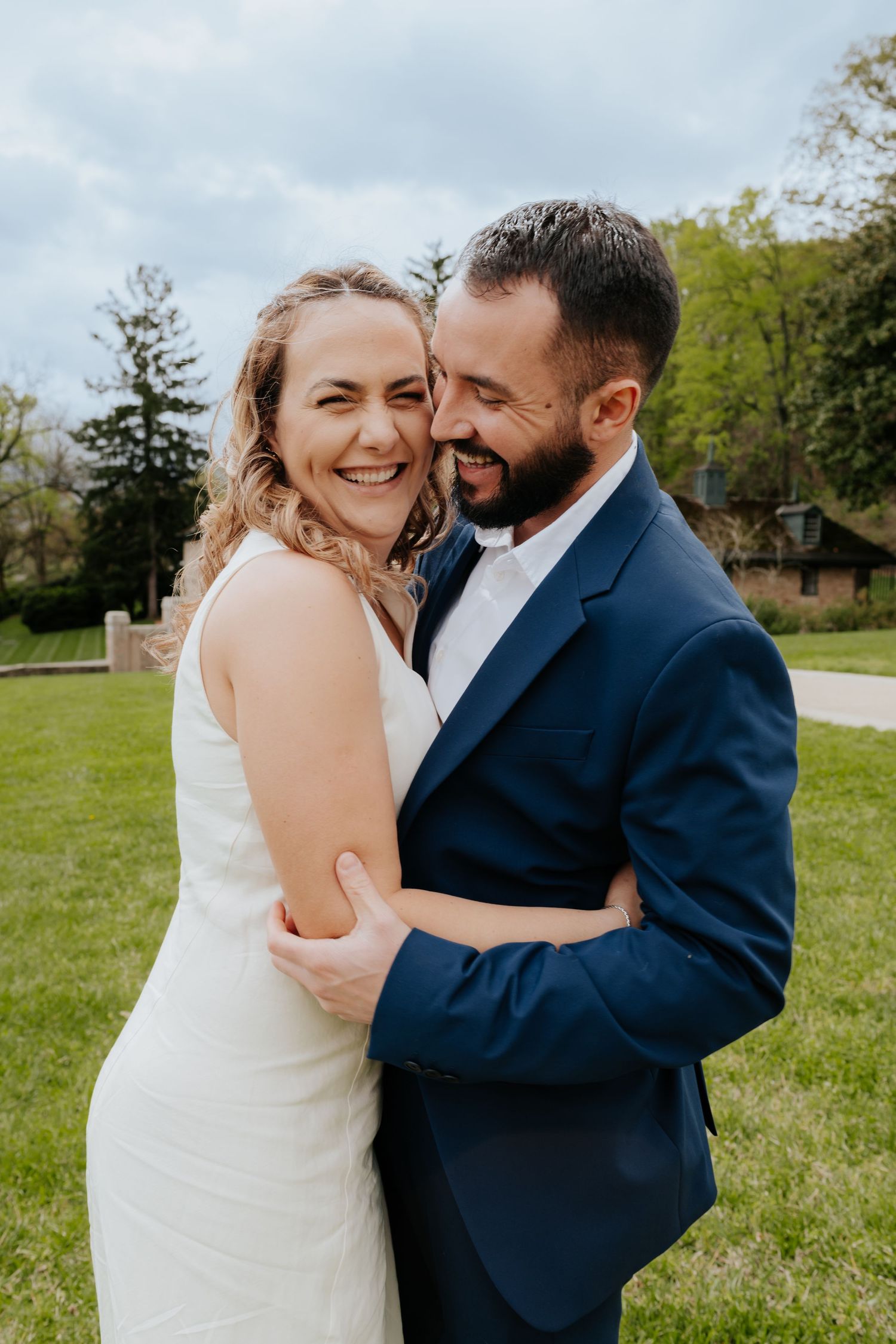 A joyful couple embraces outdoors during a wedding, with the bride in white and groom in navy blue suit on a spring day.