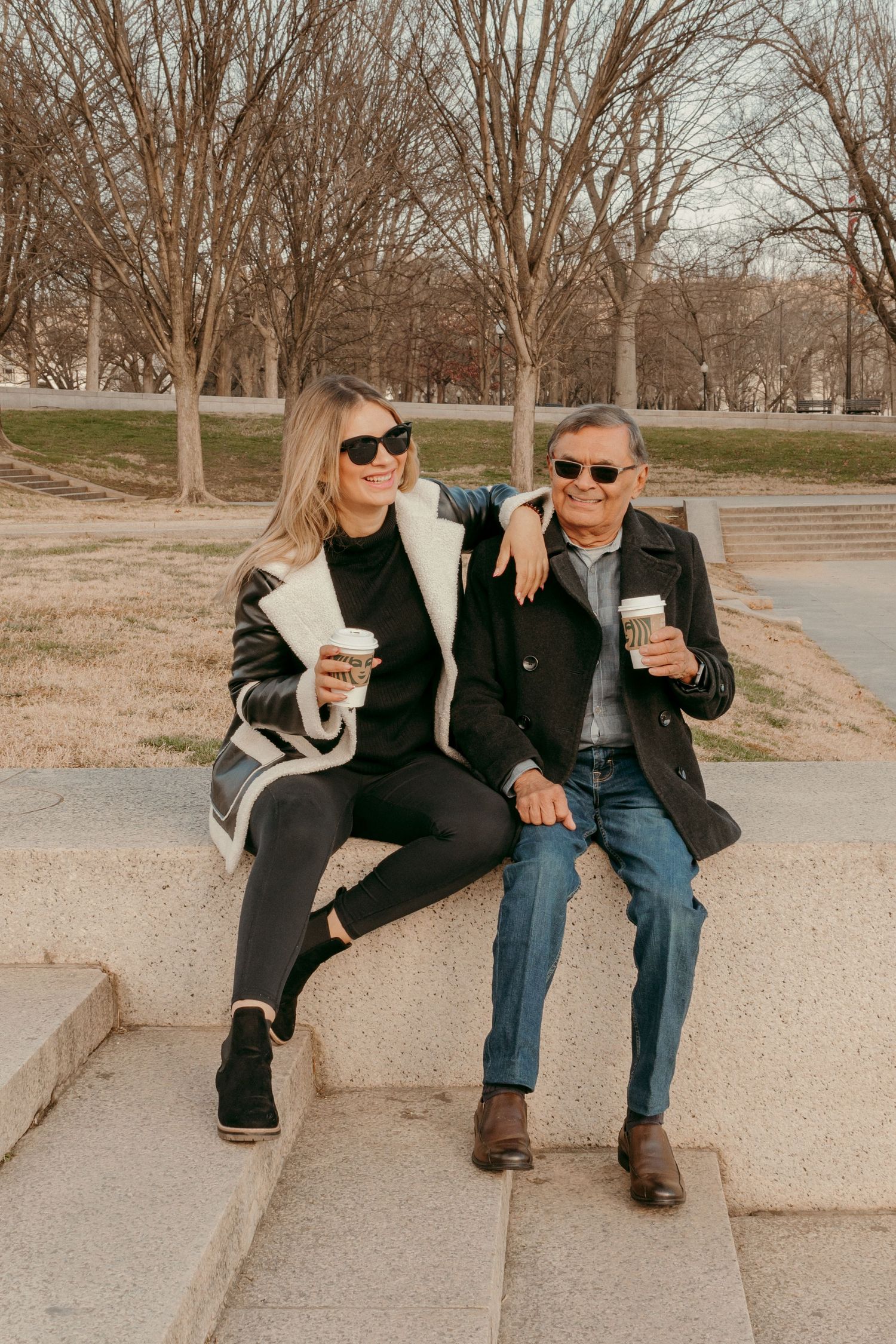 Two people in sunglasses share coffee while sitting on steps in a park during autumn.