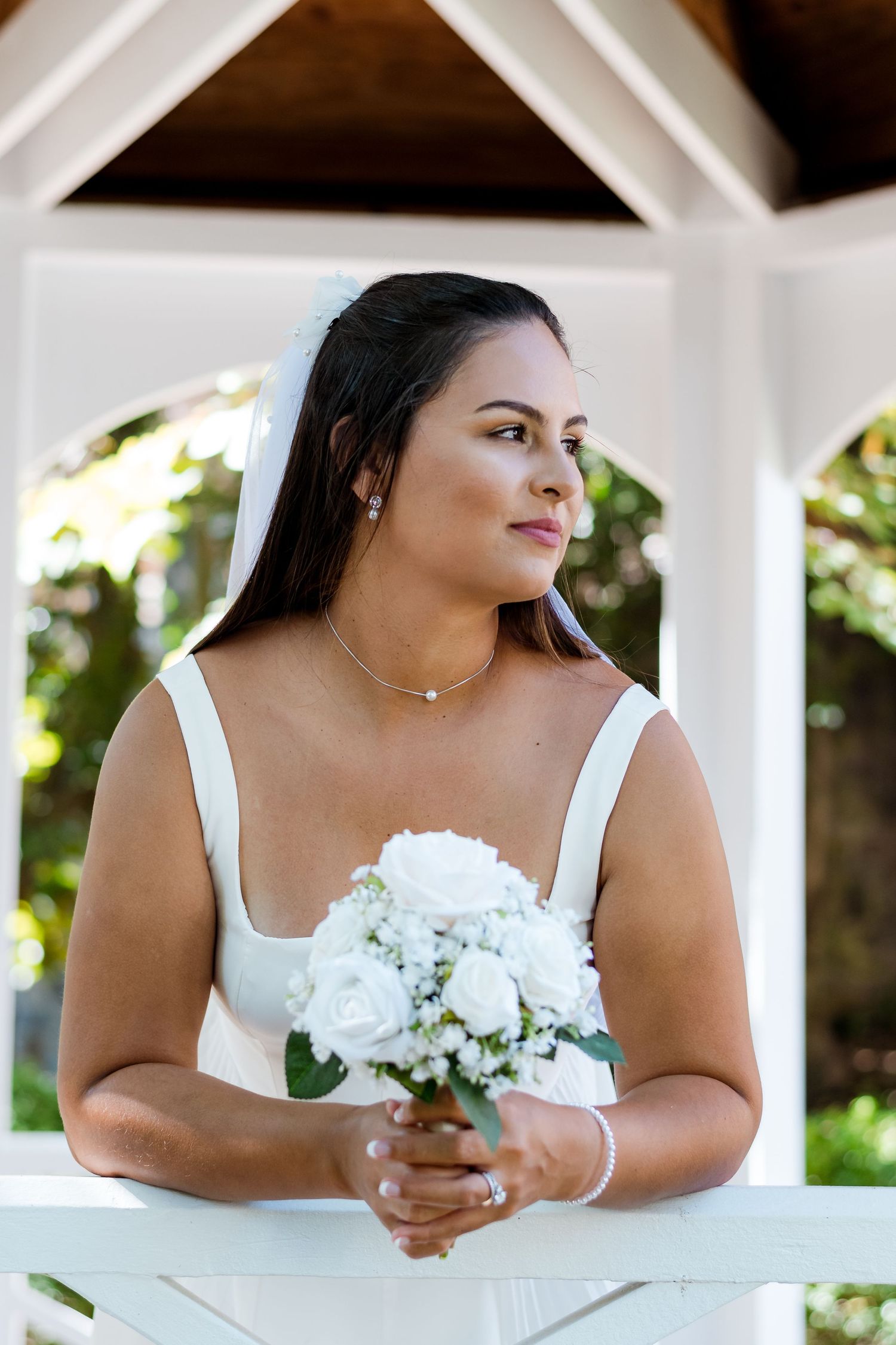 Bride in white dress holding white roses poses in elegant gazebo on wedding day.