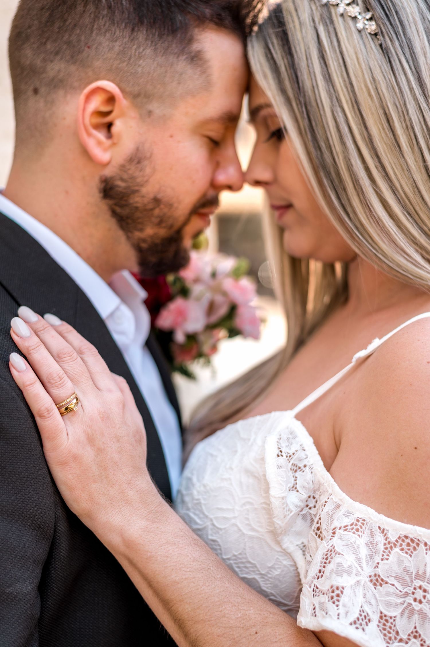 A romantic wedding portrait showing a close-up tender moment with pink flowers and lace dress details.