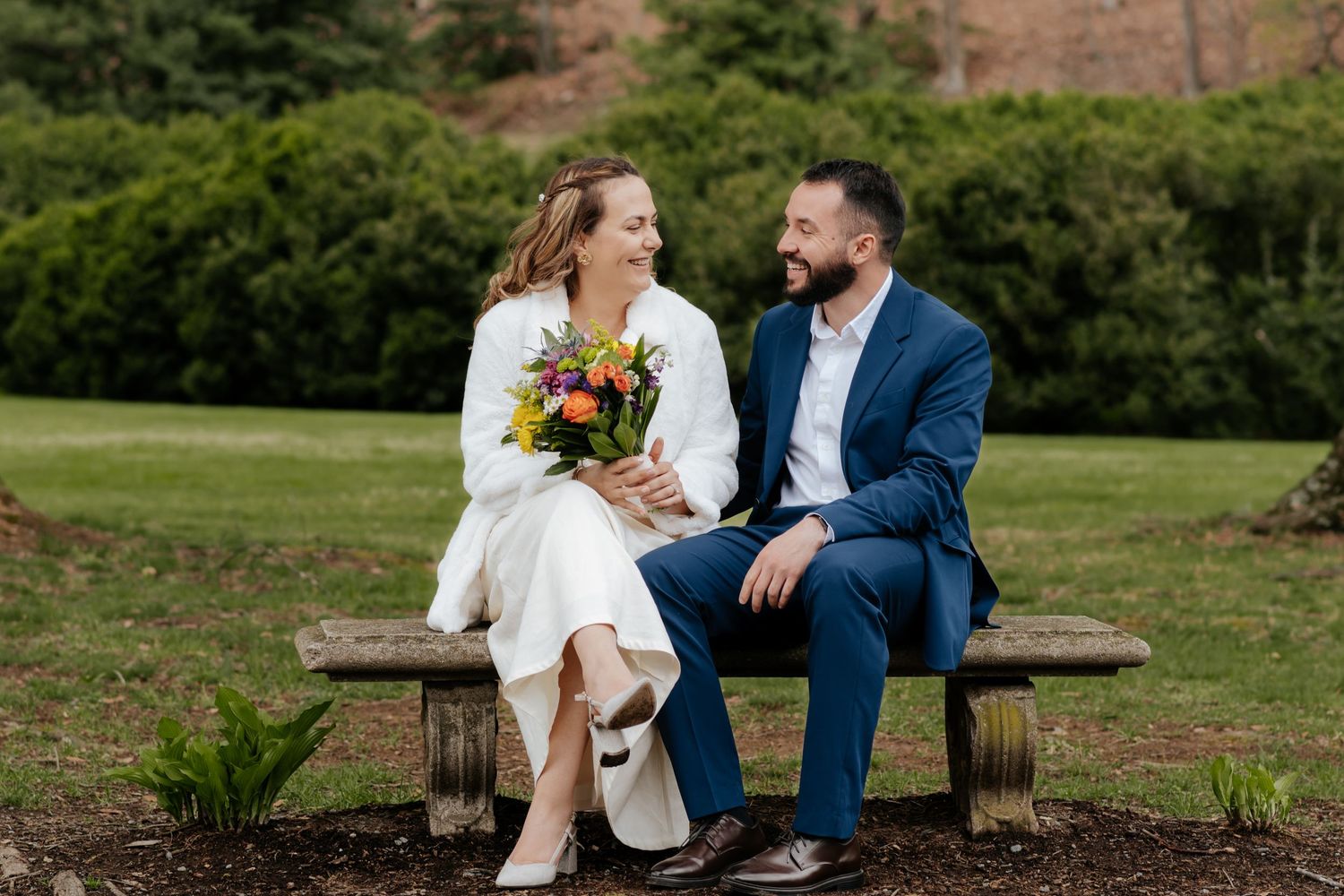 A couple sitting on a wooden bench outdoors share a joyful moment surrounded by greenery while holding a sunflower bouquet.