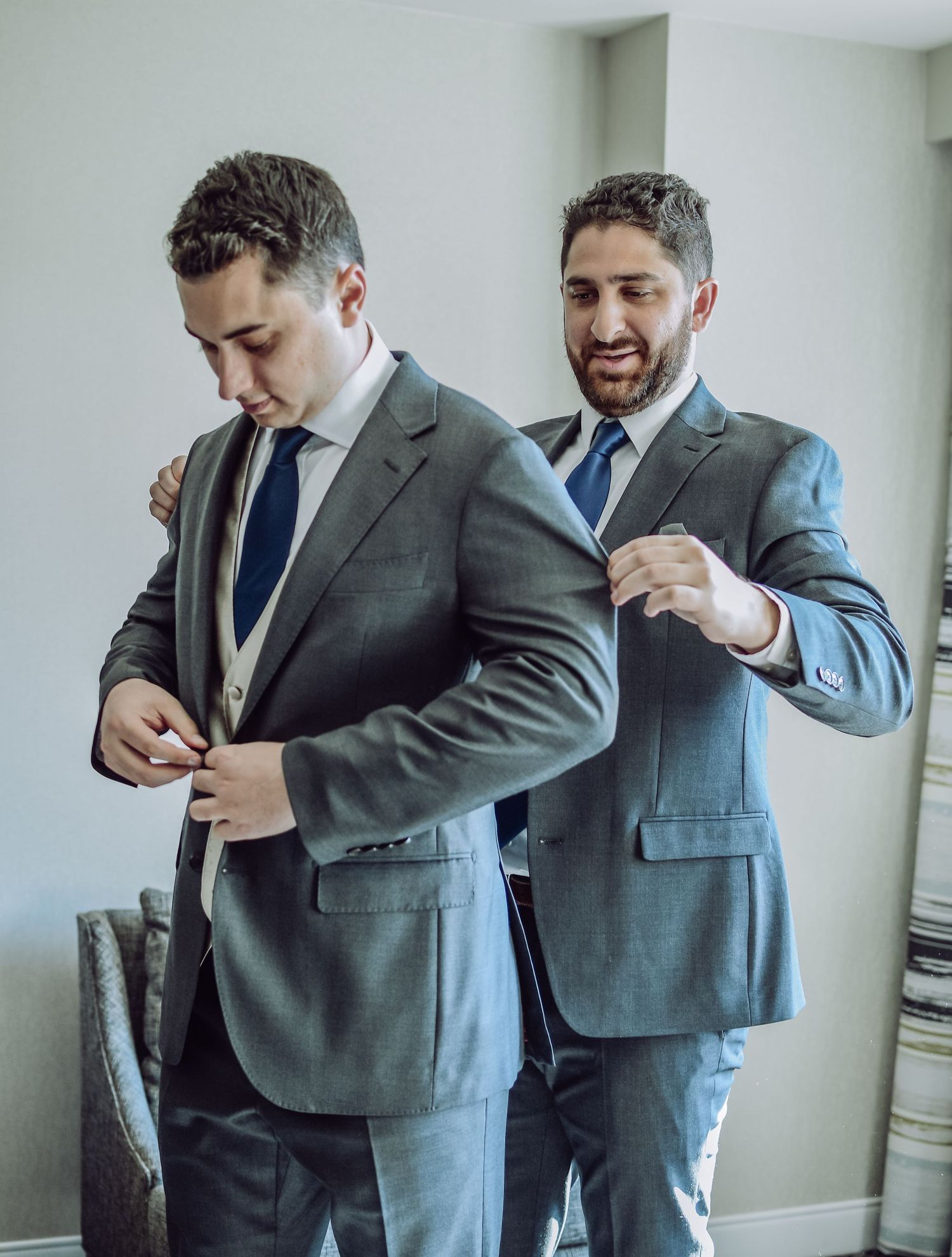 Two men in gray suits and navy ties help each other get dressed and adjust their formal attire for a special occasion.