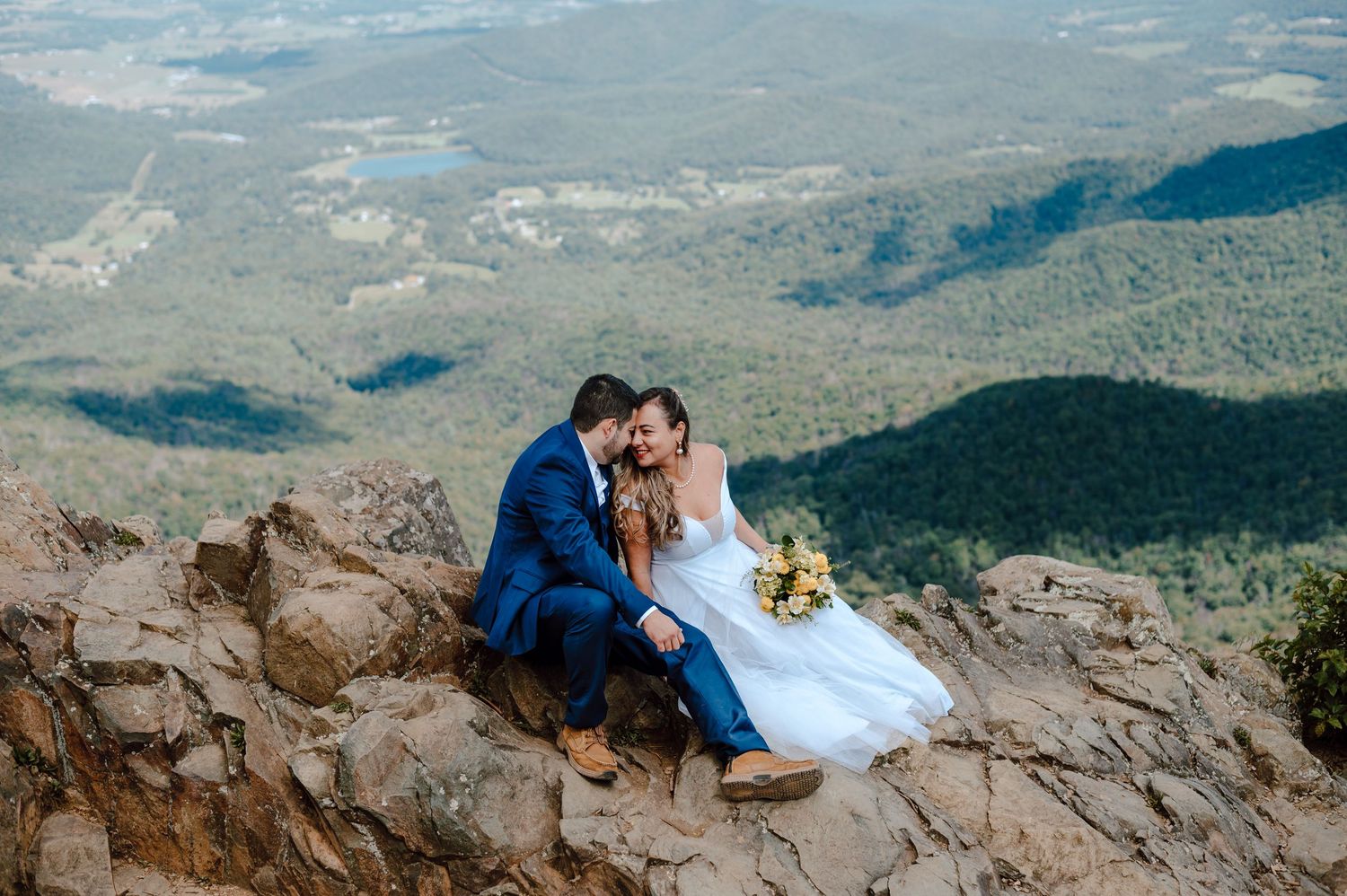 A couple shares an intimate moment on a rocky mountain overlook with a vast landscape view stretching to the horizon.