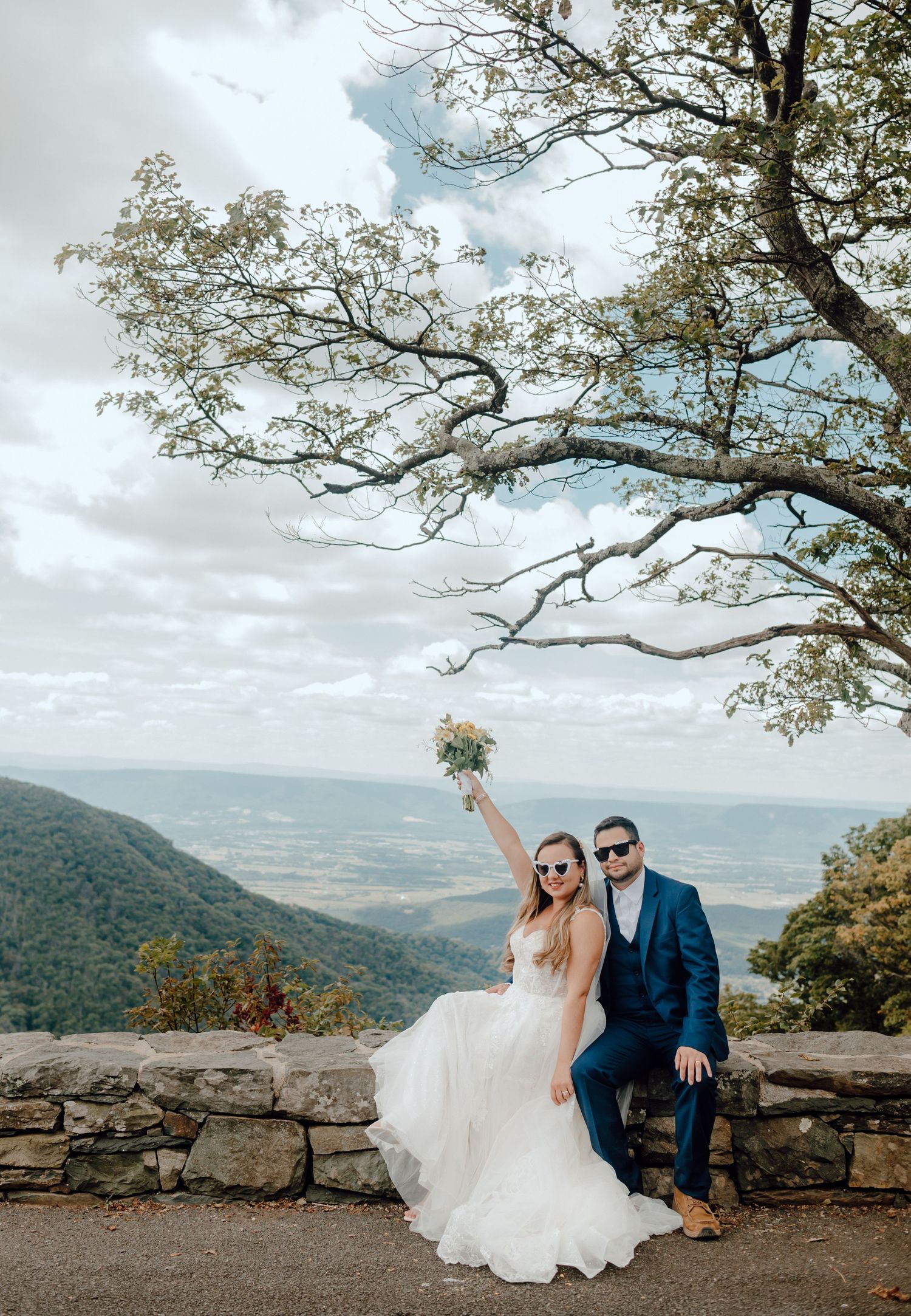 A couple poses on a scenic mountain overlook with dramatic cloudy skies and tree branches framing the vista.