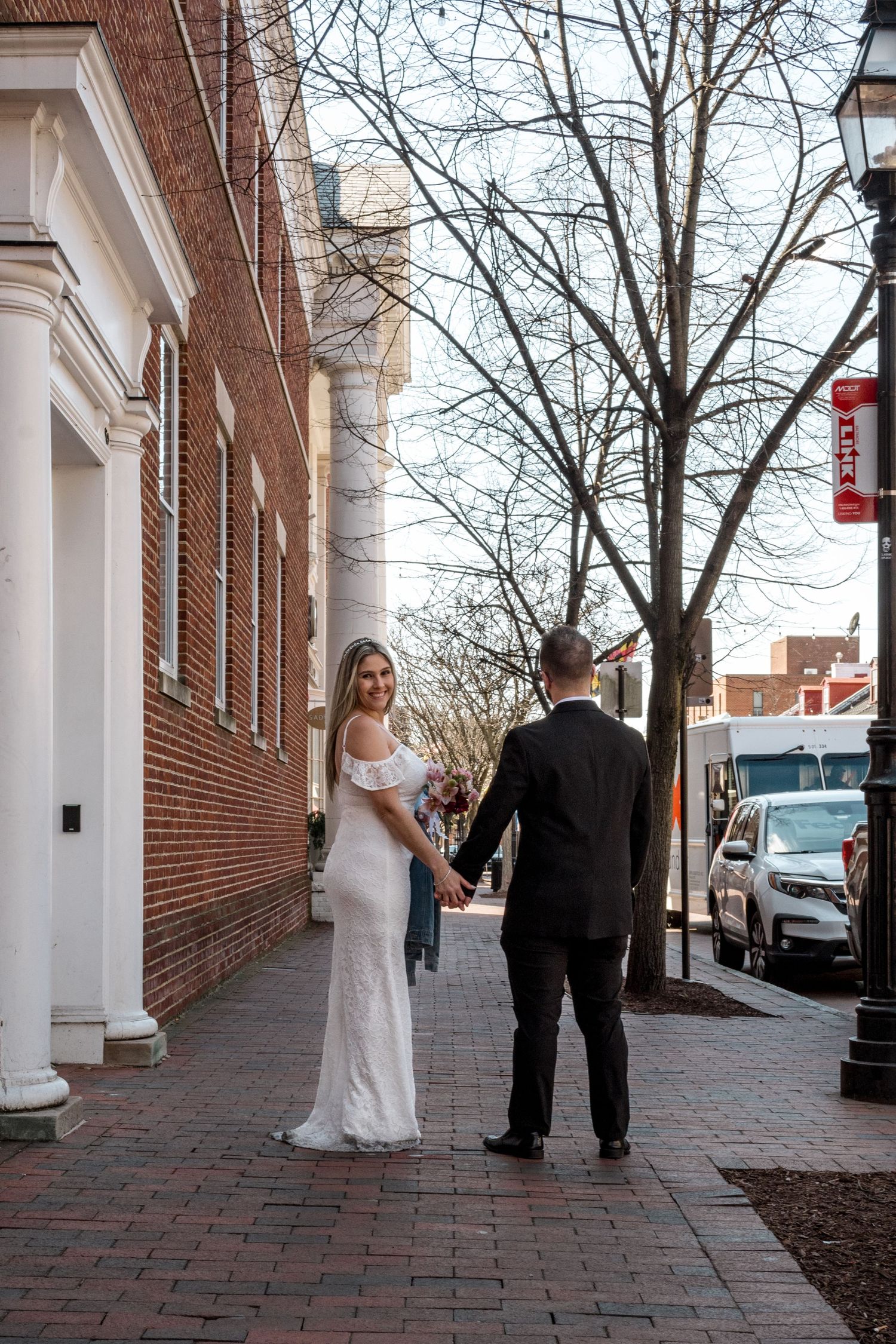 Elegant wedding couple walking hand in hand on brick sidewalk by historic colonial building.