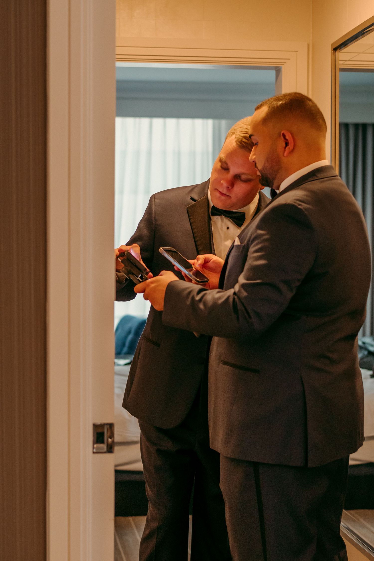 Two people in suits look at their phones together in a warmly lit interior room.