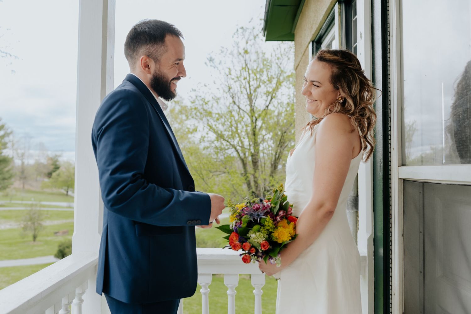 The bride and groom share a joyful moment on a white porch while holding a vibrant orange and yellow bouquet.