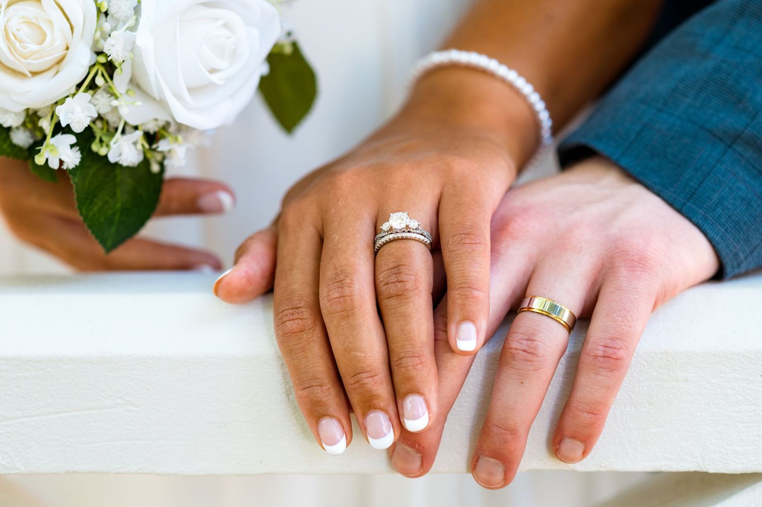 Wedding rings displayed on couple's hands resting on white railing with bouquet in background.