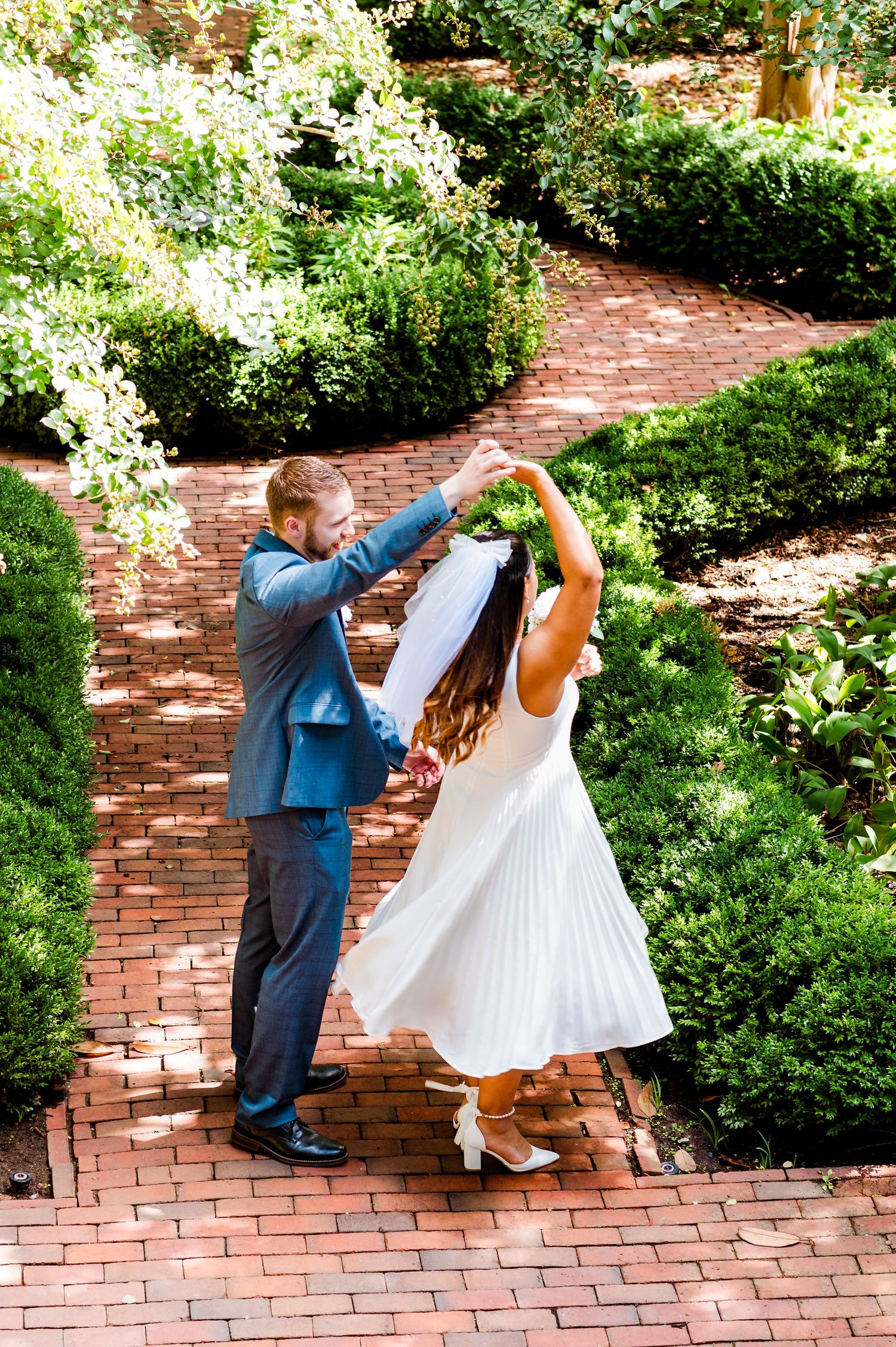 Playful wedding dance on brick pathway surrounded by manicured garden hedges.