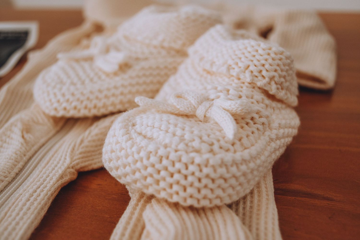 Handmade knitted off-white baby booties with bows displayed on a wooden surface next to a cable knit blanket.