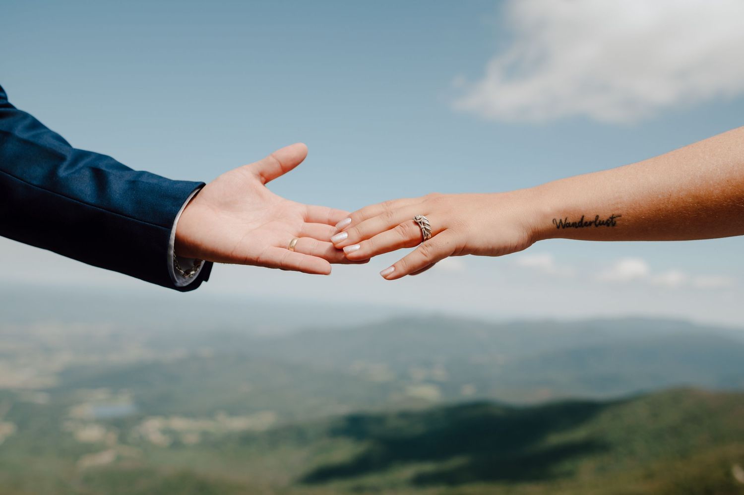 Two hands reaching across with one hand showing an engagement ring against a cloudy outdoor mountain backdrop.