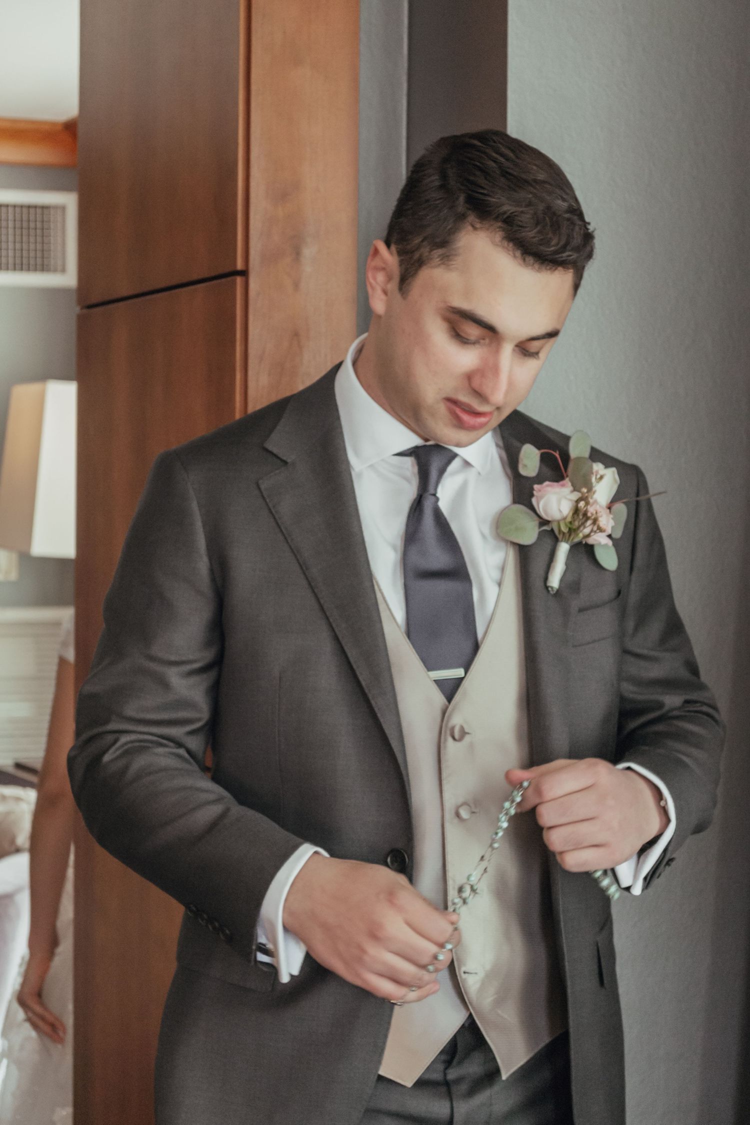 A groom in a charcoal suit adjusts his boutonniere and vest before the wedding ceremony.