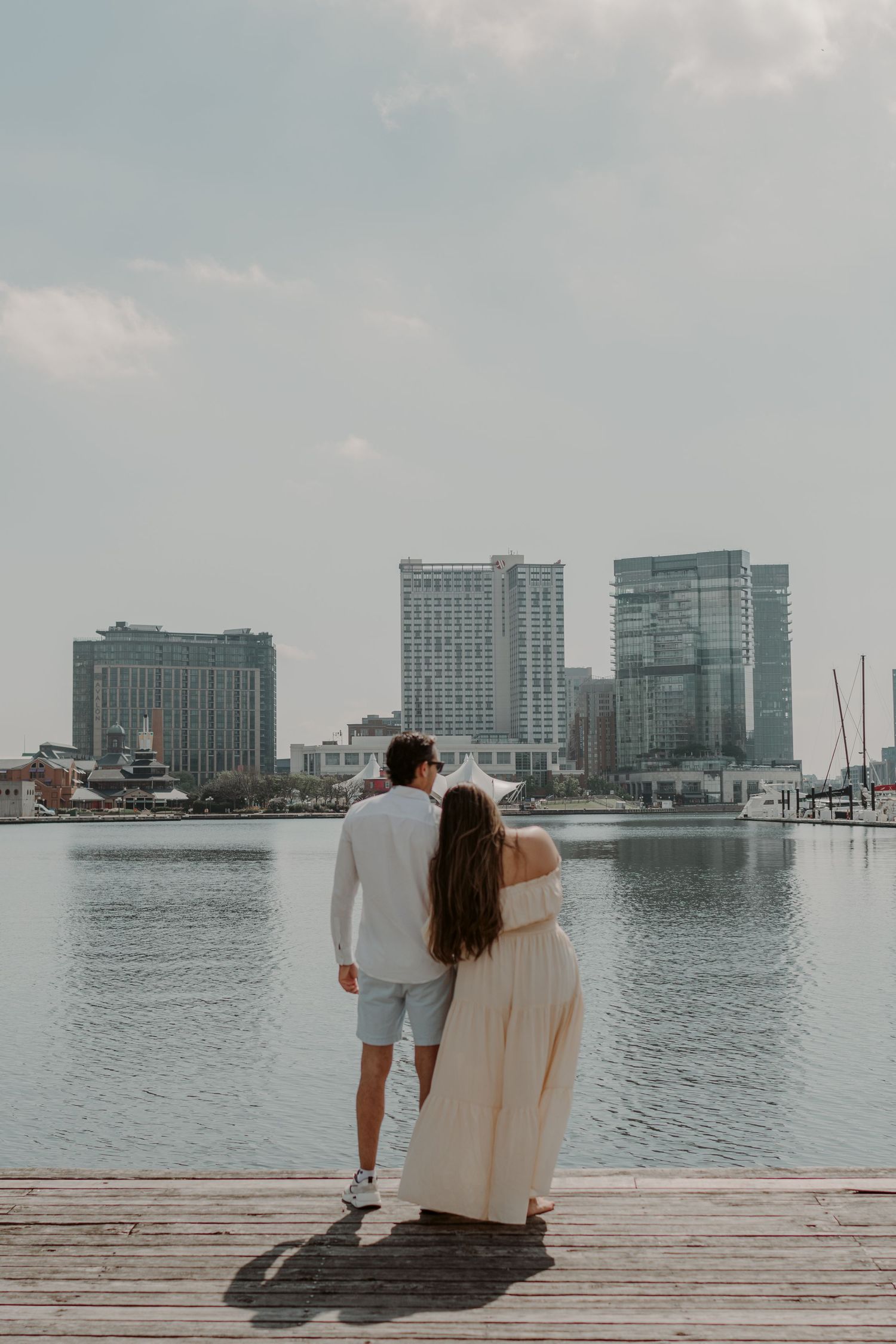 Couple standing on wooden pier overlooking Baltimore Inner Harbor with city skyline and high-rise buildings in background.