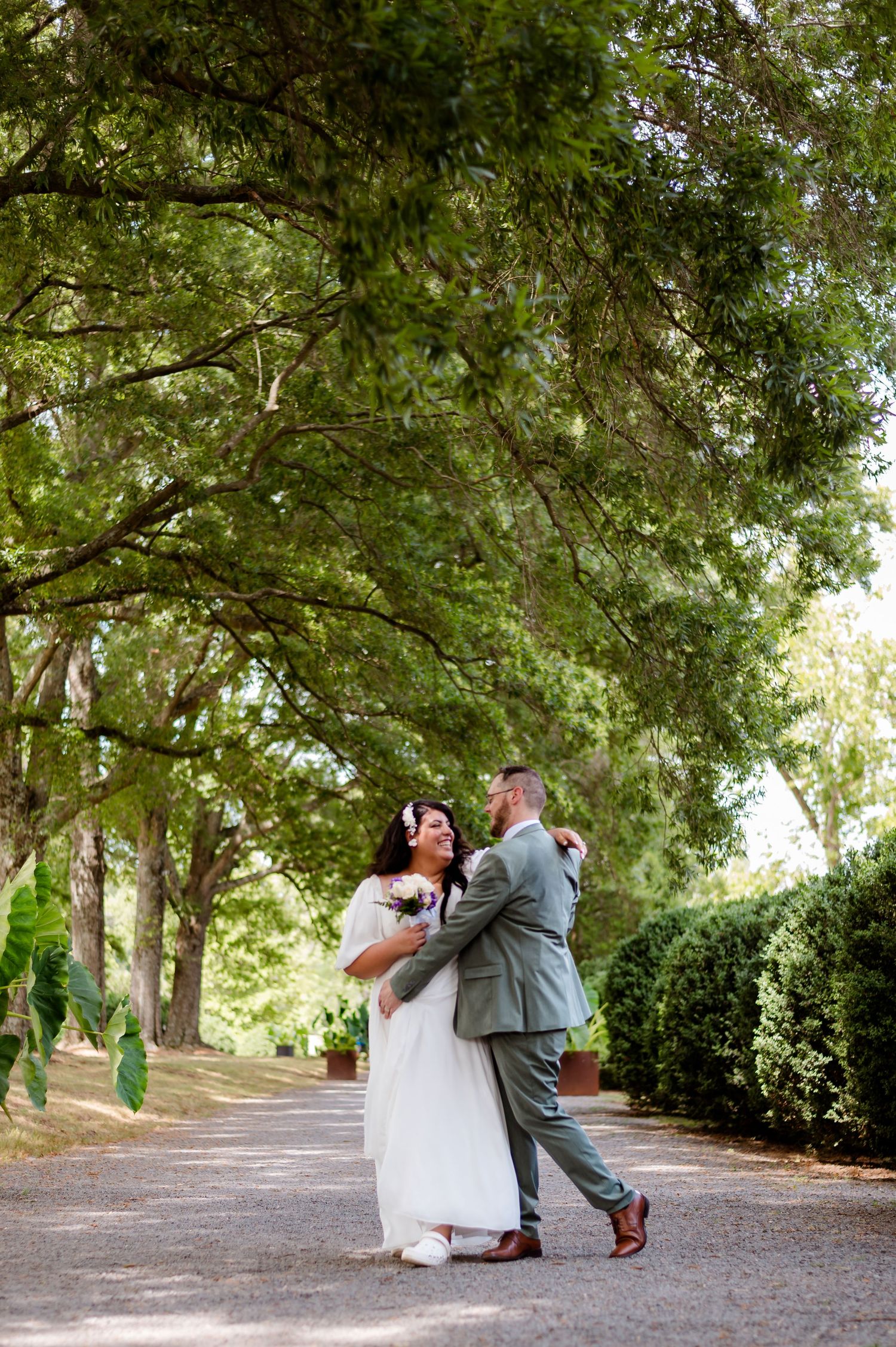 A romantic couple embraces on a tree-lined pathway in an outdoor garden wedding photoshoot.