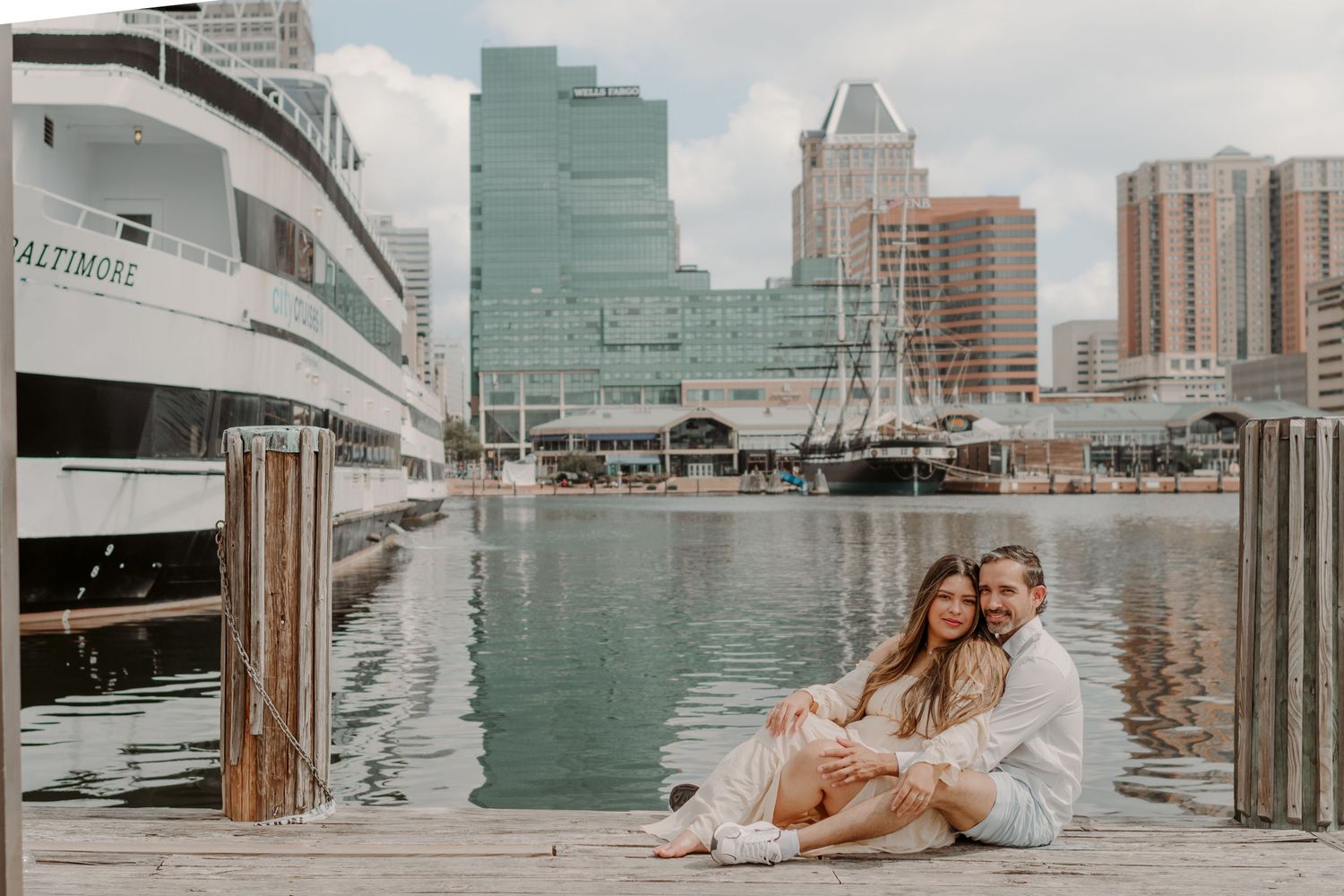 A couple sits romantically on a wooden pier overlooking Baltimore's Inner Harbor with boats and tall buildings in the background.
