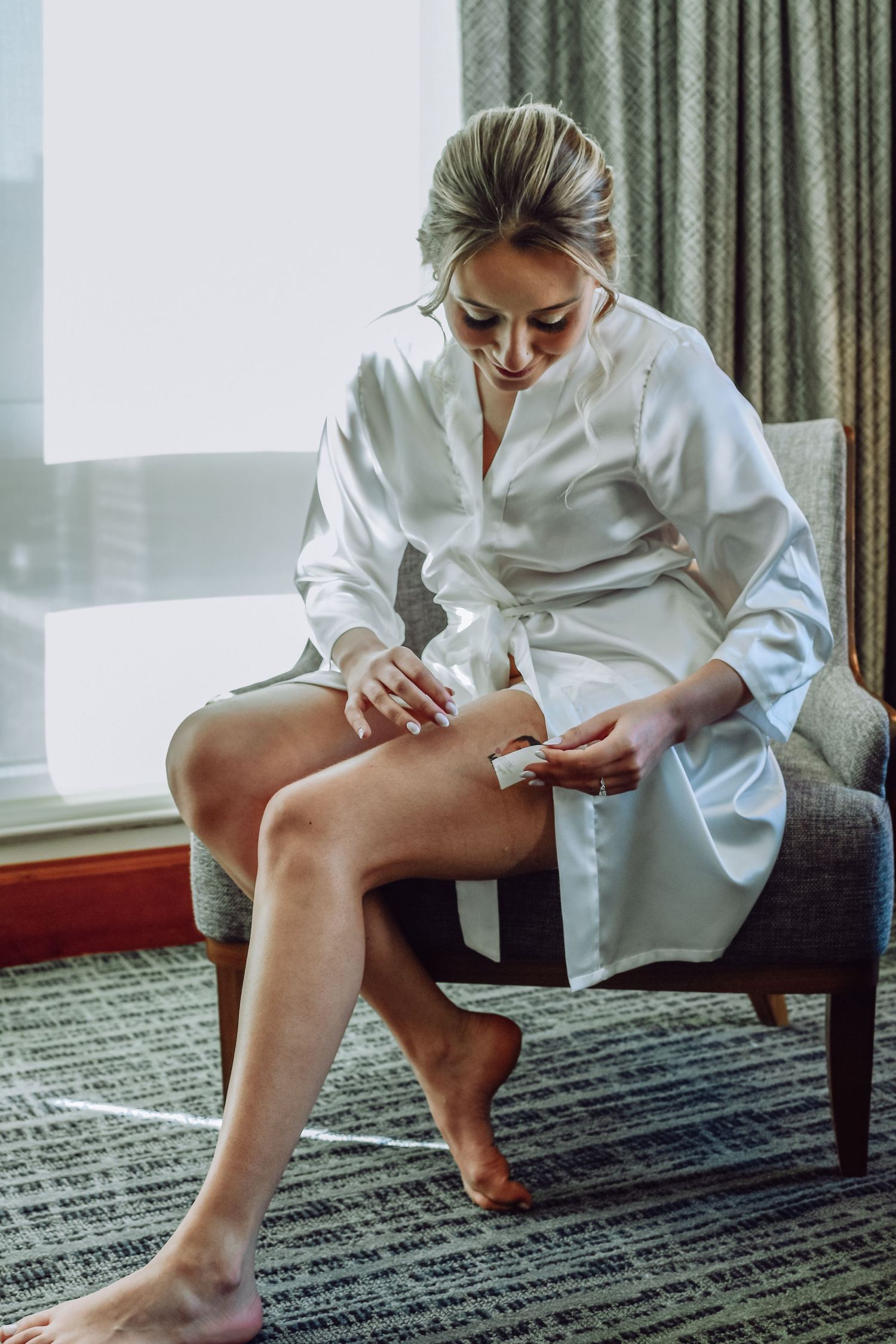 Person in white silk robe sitting on grey chair applies moisturizer to legs in sunlit modern room.