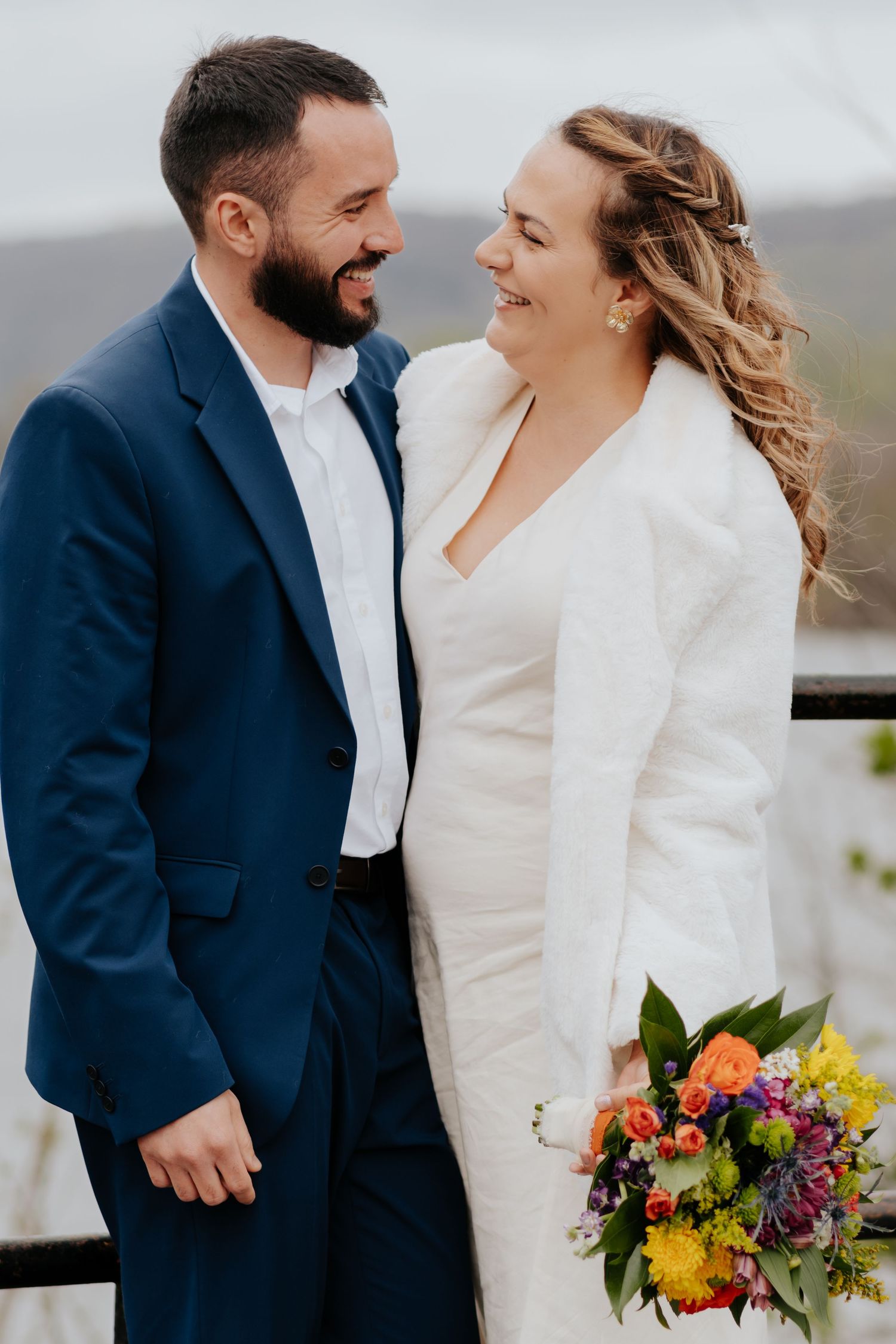 A joyful wedding couple share a laugh while wearing a navy suit and white dress with a vibrant autumn bouquet.