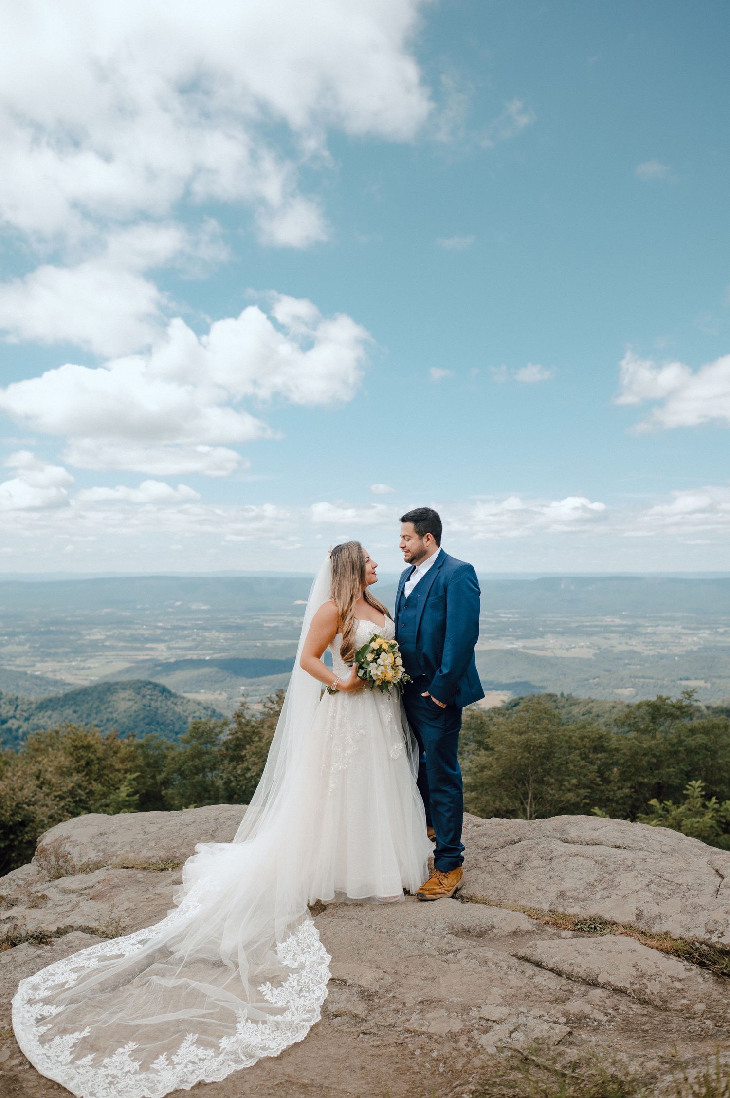 Newlyweds stand on a mountain overlook with dramatic white clouds and sweeping valley views in the background.