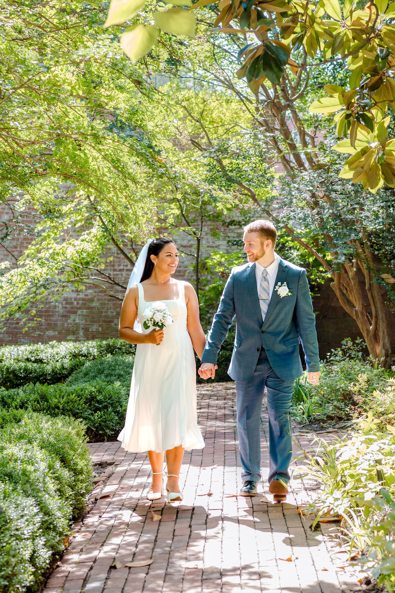 Newlyweds walking hand in hand along garden path in sunlit courtyard.