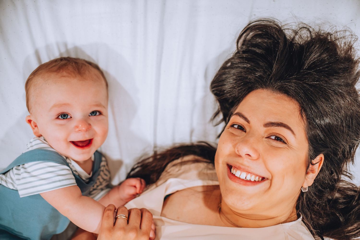 Candid lifestyle photo of parent and baby sharing a happy moment while lying on white bedding.