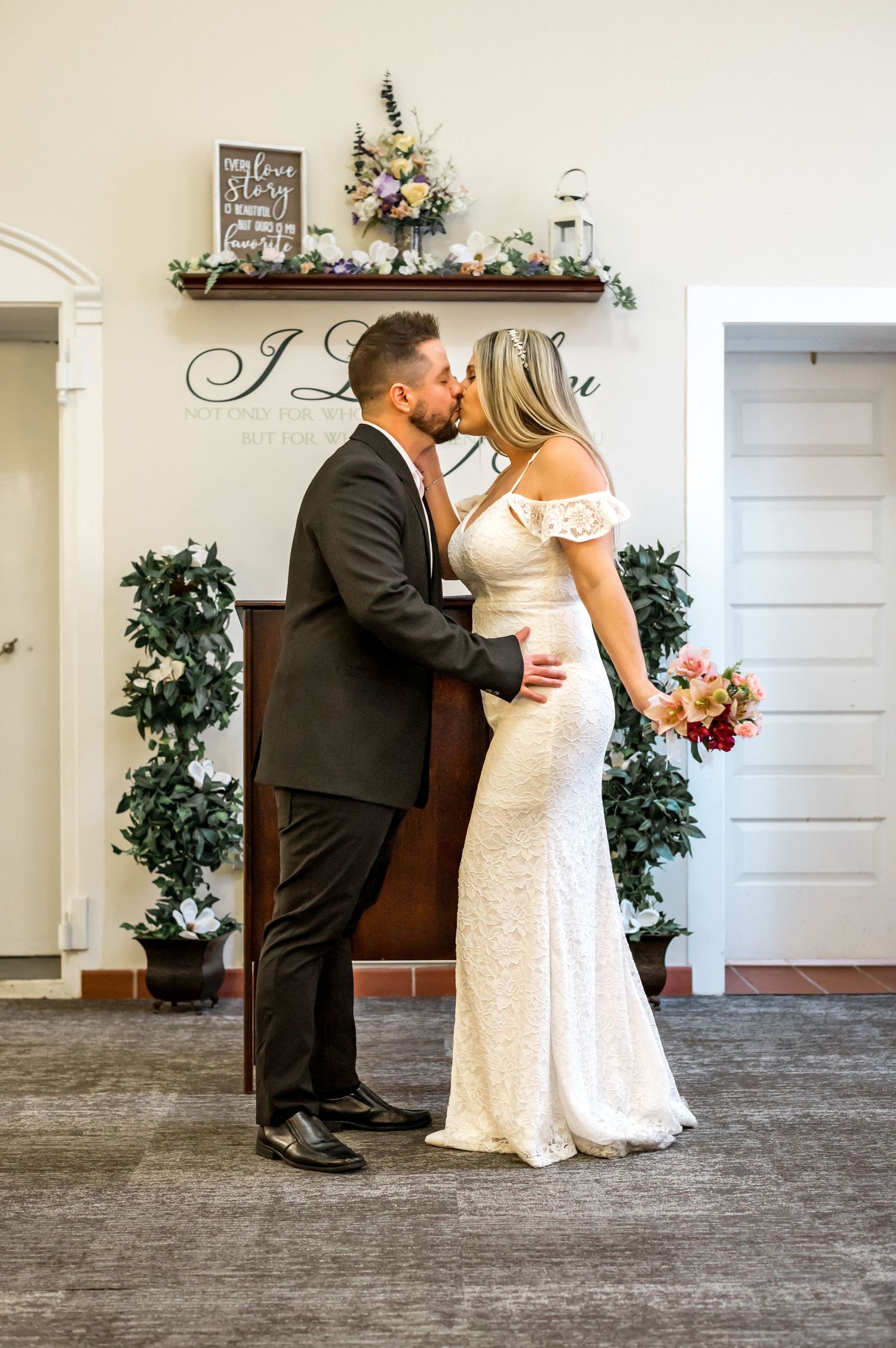 A couple shares a romantic moment under an 'I Love You' sign decorated with floral arrangements.